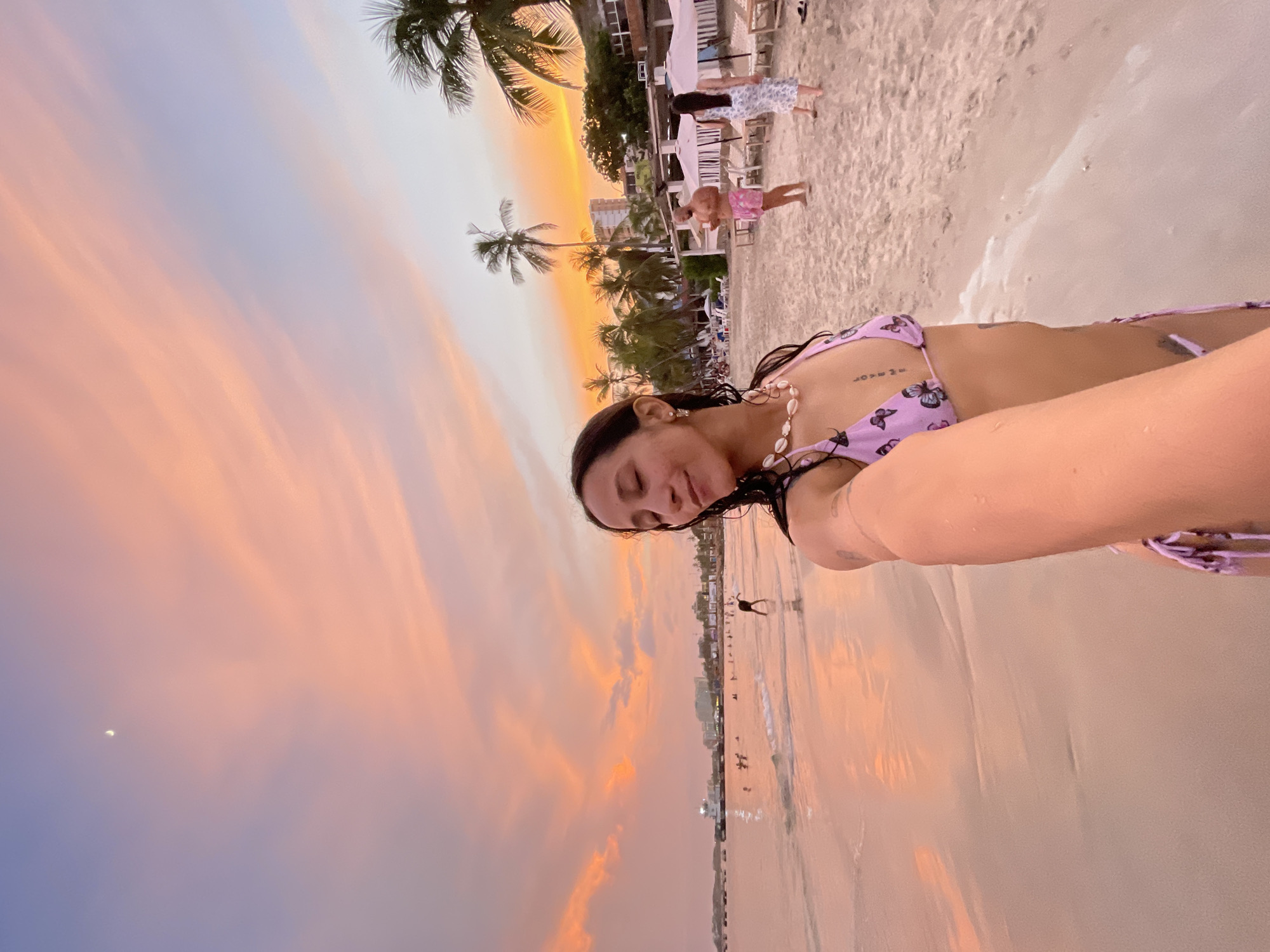 A selfie taken at beach in Pampatar, Venezuela shows a woman in a purple bikini with butterfly print, with a pearl necklace. She is holding the camera with her right hand, partially obscuring her face. Her eyes are closed, and she is smiling slightly. The background is a beach scene at sunset. The sky is a mix of orange, pink, and purple clouds. There are palm trees and buildings visible in the distance. Several people are seen on the beach, some are walking along the shore, and some are in the shallow water. A boat is visible in the water. The sand is light in color.