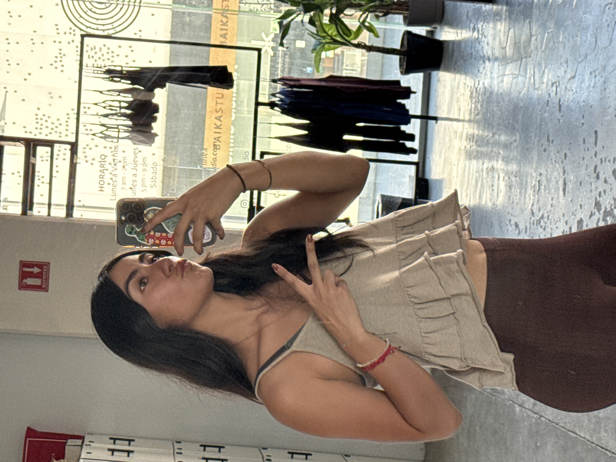 The image shows a woman taking a selfie in front of a storefront. She is holding her phone in her left hand and making a peace sign with her right. She is wearing a tank top and shorts, and her hair is dark. The storefront behind her is a clothing store called "BAIKASTU", located in the city of Puebla, Mexico. A sign on the window indicates the store hours. There are clothes hanging on racks inside the store, and plants in pots on the window sill. The floor appears to be concrete. A red extinguisher sign is mounted on the wall.