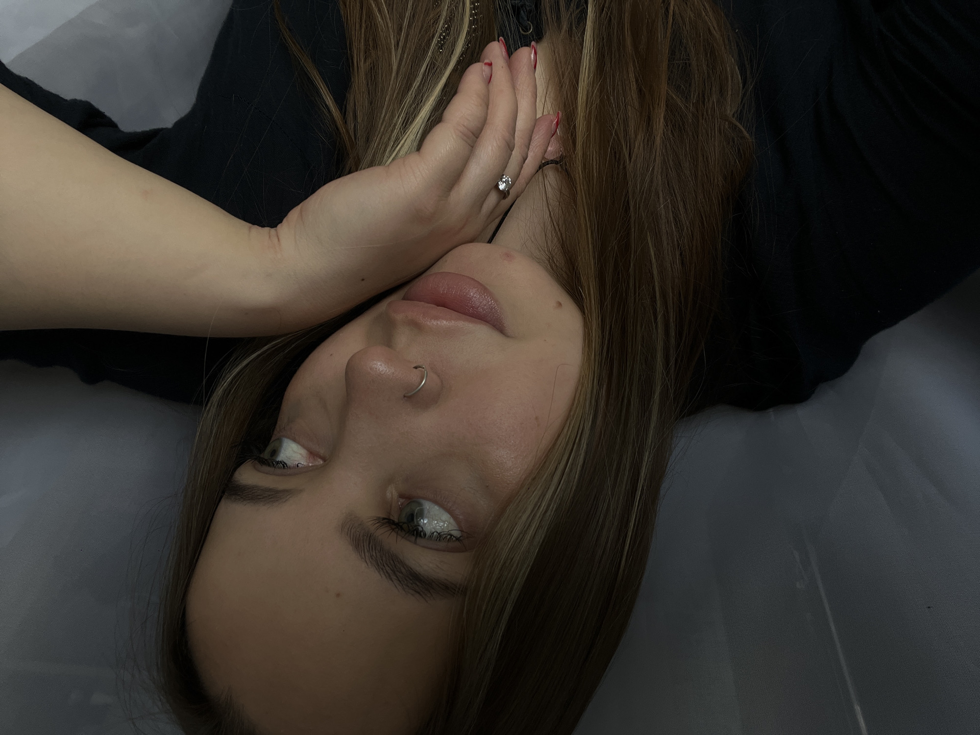 A young woman is lying down, possibly on a gray or white surface, with her head oriented to the lower left of the image. She has fair skin, light brown hair with blonde highlights, and light-colored eyes. A small ring is visible on one of the fingers of her left hand, which is positioned near her face. She has a silver nose ring in her left nostril. The background includes black clothing and an indeterminate surface. The location of Novopokrovskaya, Russia, cannot be determined from the image.