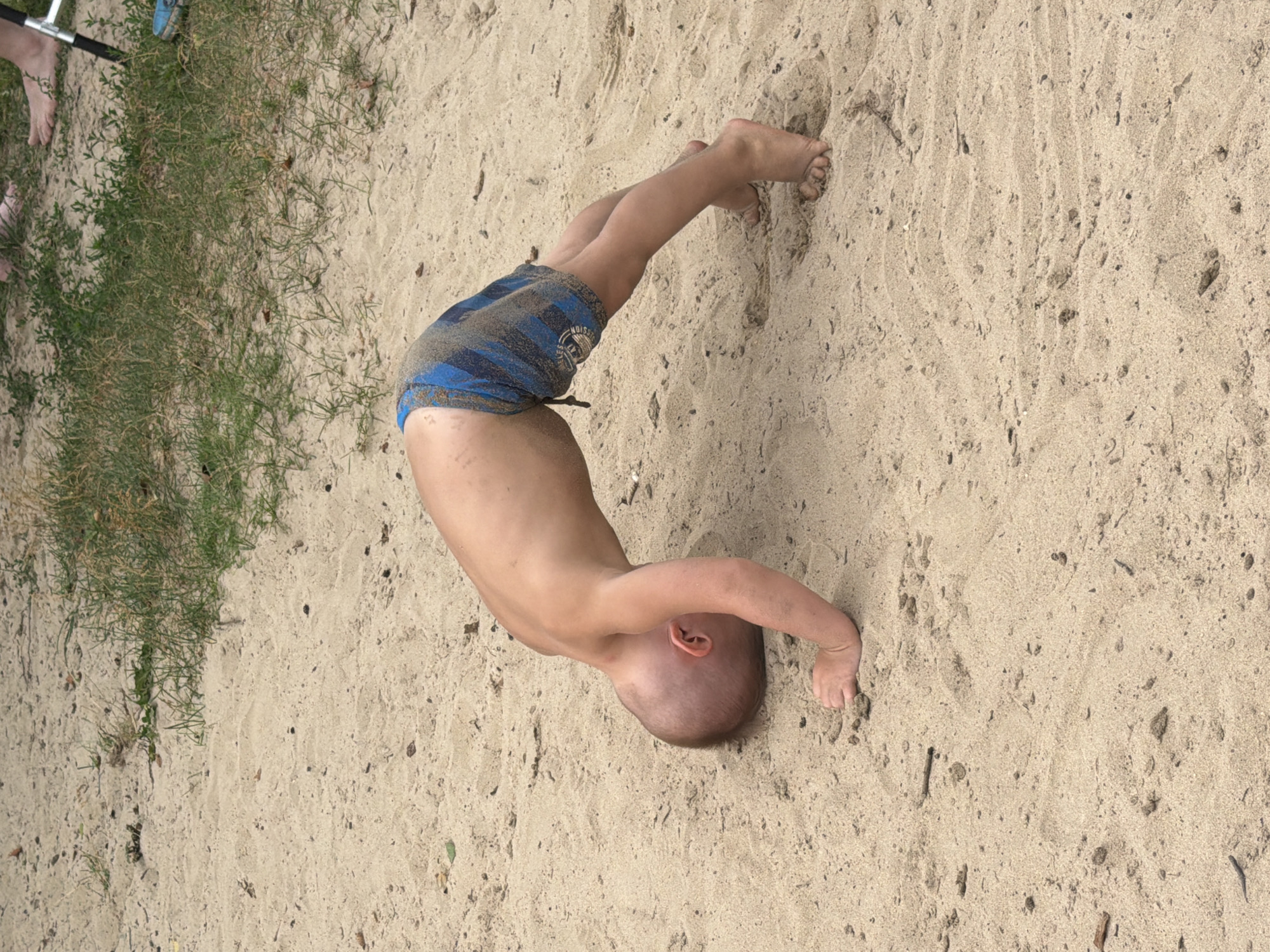 A young child with a shaved head is doing a handstand or backbend on a sandy beach. The child is wearing blue and black striped swim shorts and their body is arched. Their hands are on the sand, and their feet are in the air. The beach is covered in light brown sand with some small pebbles and debris. To the left of the child, there is a patch of green grass with some dry blades. In the upper left corner, a person's feet and lower legs are visible, wearing a blue shoe and showing bare skin. There is also a metal pole with black handles, possibly part of a recreational item. The scene appears to be outdoors during the daytime, with natural light illuminating the sand and child. The child's expression is not visible, but their posture suggests activity and play. There is a small, circular logo on the swim shorts with some text, but it is not legible. The setting is a sandy area that could be a beach or a playground. The location is identified as Malakhovka, Russia.