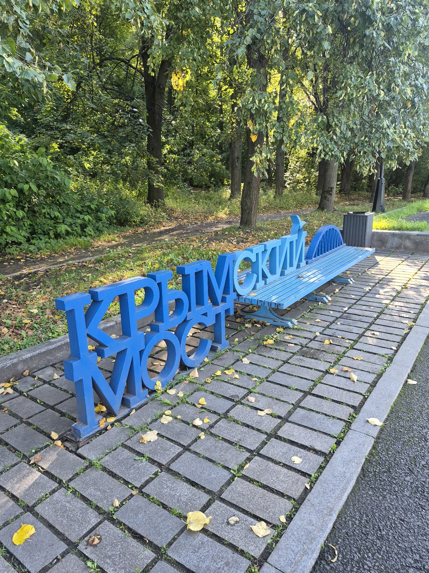 The image depicts a blue bench with Cyrillic lettering spelling "КРЫМСКИЙ МОСТ" (Crimean Bridge) in large, three-dimensional letters. The bench itself is also blue and has a curved backrest. The lettering is positioned in front of and partially integrated into the bench. The scene is set outdoors in a park-like environment with trees and foliage in the background. The ground is paved with grey rectangular stones, and fallen yellow leaves are scattered across the paving and the grass. The lighting suggests it is daytime, with sunlight filtering through the trees. To the right of the bench, a dark grey trash bin is visible, and beyond that, a grassy area and a path. This installation is located in Moscow, Russia, specifically at the Muzeon Park of Arts, near the Krymsky Bridge. There are no people visible in the image.