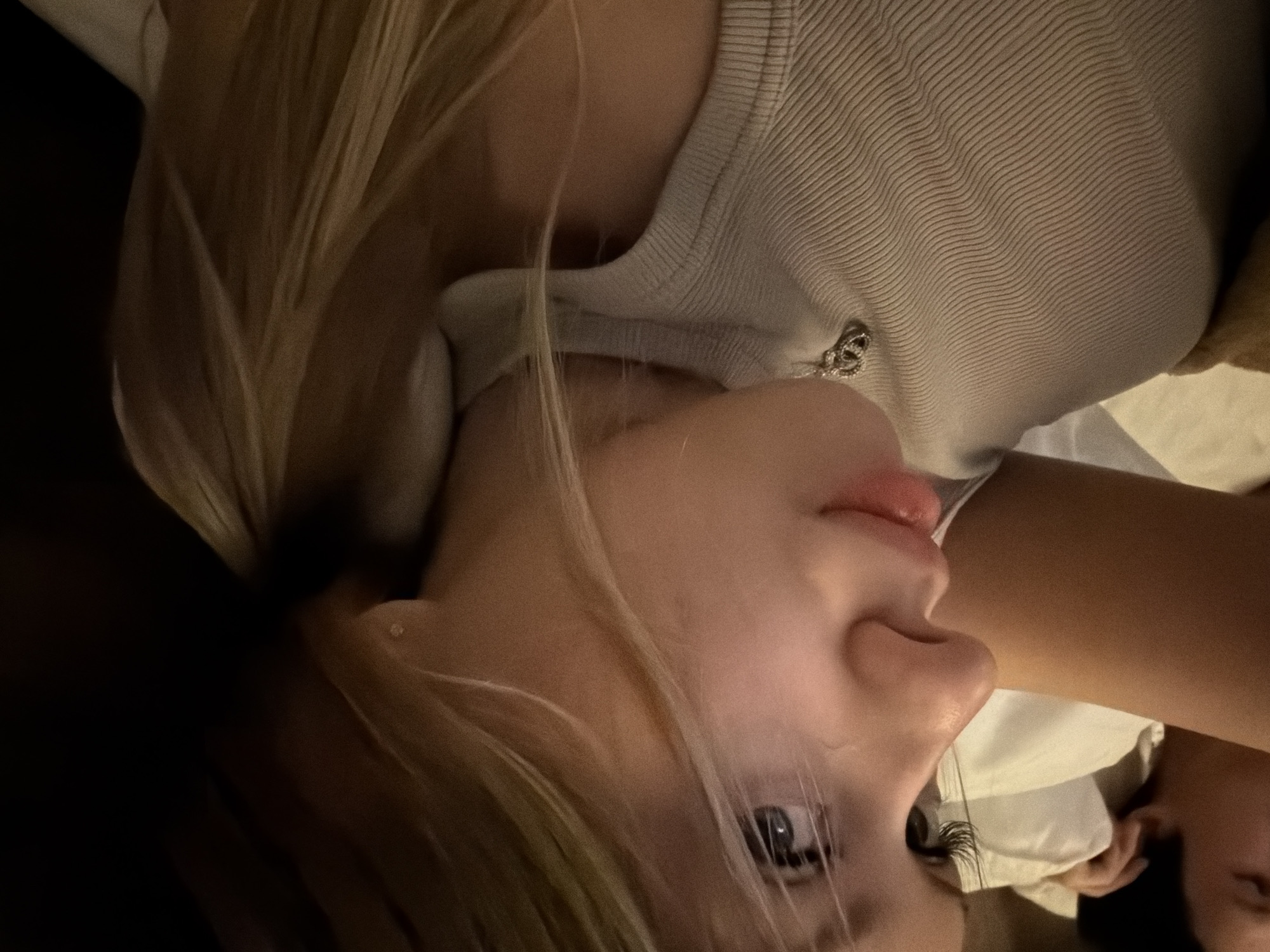 The image displays a close-up, profile view of a young woman with blonde hair, wearing a ribbed, light-colored sleeveless top and a silver necklace. Her head is tilted, and her gaze is directed to the left, with her eyes partially visible. In the background, another person's shoulder and arm are visible, suggesting they are lying down together. The scene appears to be indoors, with soft lighting. There is no discernible connection to the city of Đà Nẵng, Vietnam, as no elements in the image provide geographical context.