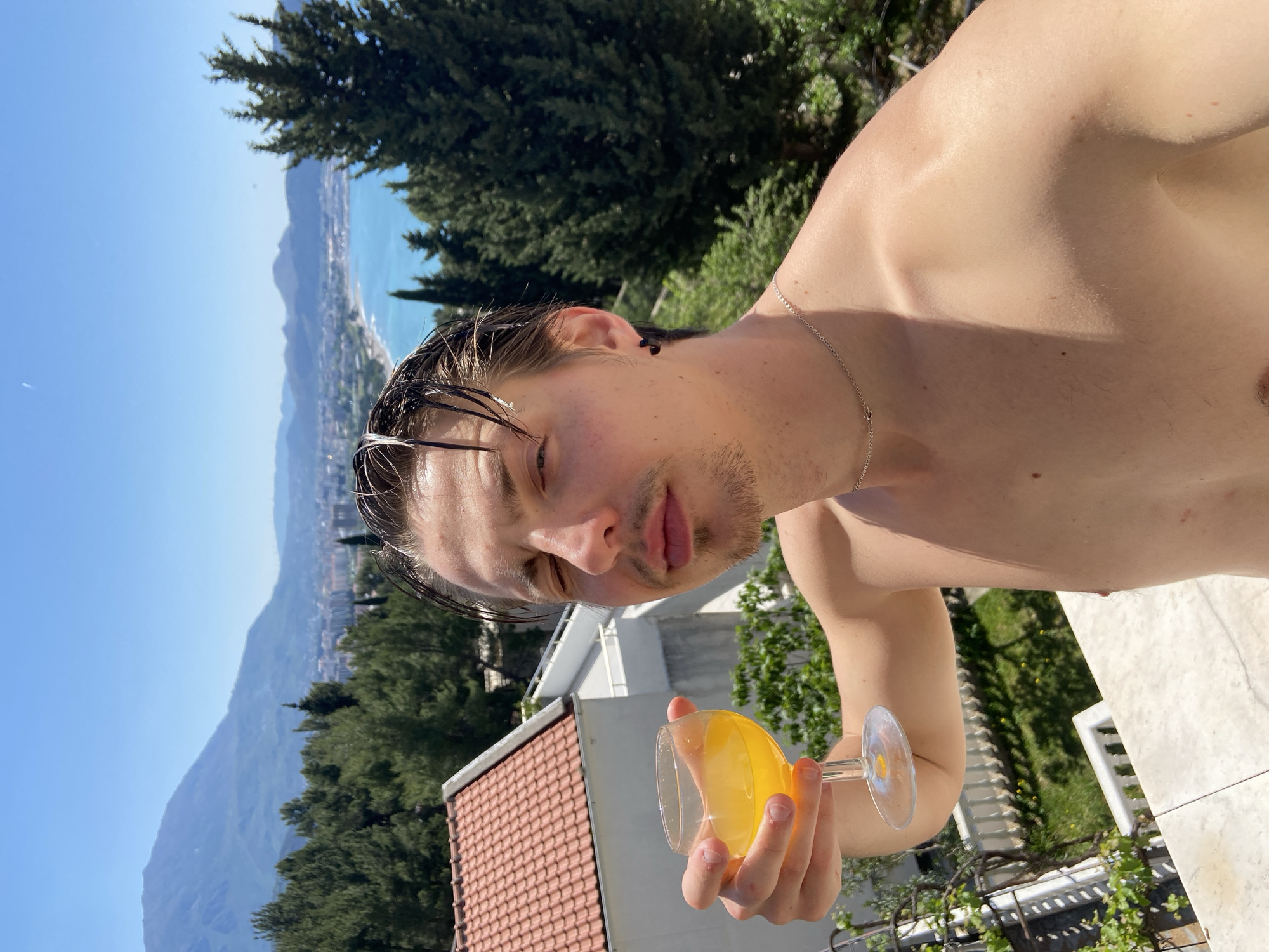 A man, bare-chested except for a thin silver necklace, is holding a stemmed glass filled with an orange liquid. The background reveals a mountainous landscape with a town nestled along the coast. The setting appears to be Šušanj, Montenegro, given the architectural style and geographical features. The man's hair is wet and swept back. A tiled roof is visible in the lower left. The angle suggests the photo was taken at an elevated position.