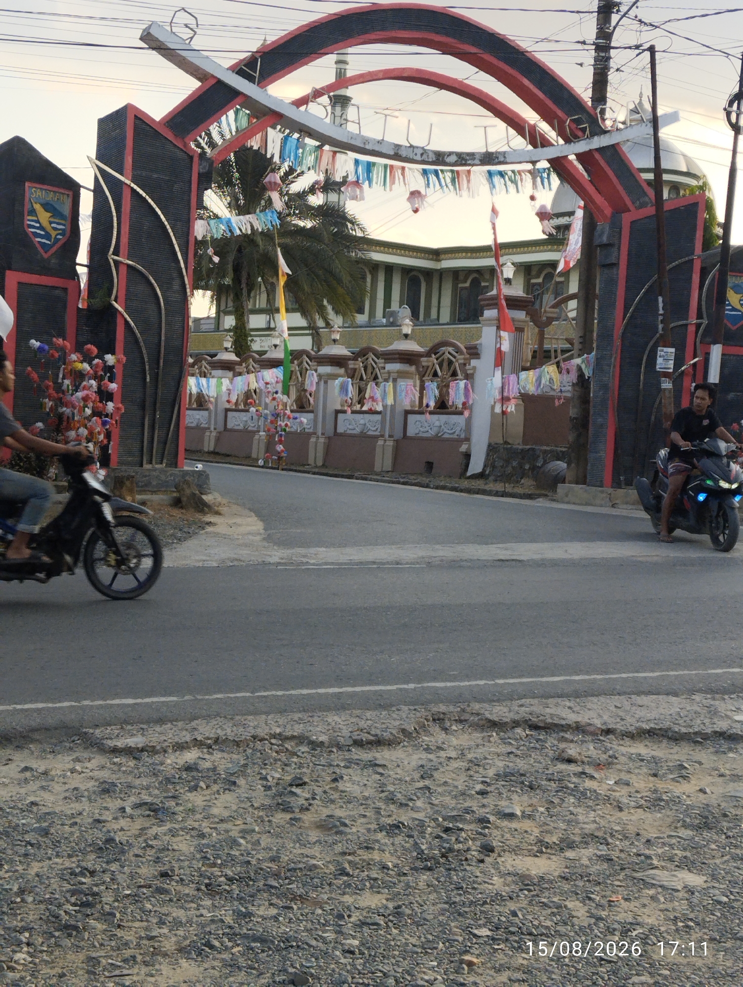 The image depicts an archway in Serongga, Indonesia, likely an entrance to a community area or religious site, given the presence of a mosque or temple in the background. The archway is decorated with colorful flags and streamers. In the foreground, two men are seen riding motorcycles on a paved road. The man on the left is partially visible and is wearing a grey shirt and blue jeans. The man on the right is wearing a black t-shirt and shorts. The road surface in the foreground is rough and appears to be under repair. The sky is overcast, suggesting late afternoon or early evening. The timestamp "15/08/2026 17:11" is visible in the bottom right corner, indicating the date and time the image was captured.
