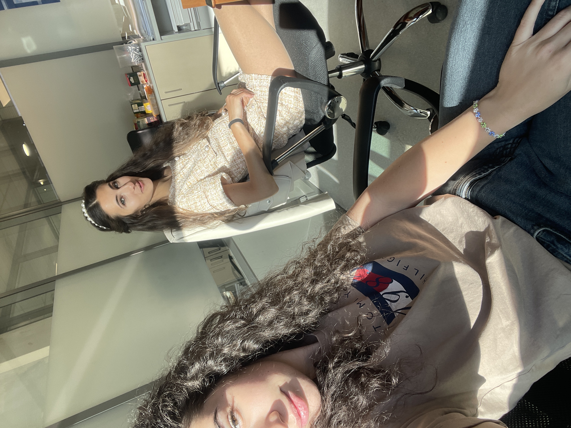 The image captures two young women in an indoor office setting. One woman, with dark curly hair and wearing a Tommy Hilfiger t-shirt, appears to be taking a selfie. The other woman, with long brown hair and wearing a light-colored dress, is sitting in an office chair in the background. There's office furniture visible, including a cabinet with items stored on shelves. The environment suggests an office interior with bright natural light entering from a window. There is no indication of the location of the city of Sofia, Bulgaria.