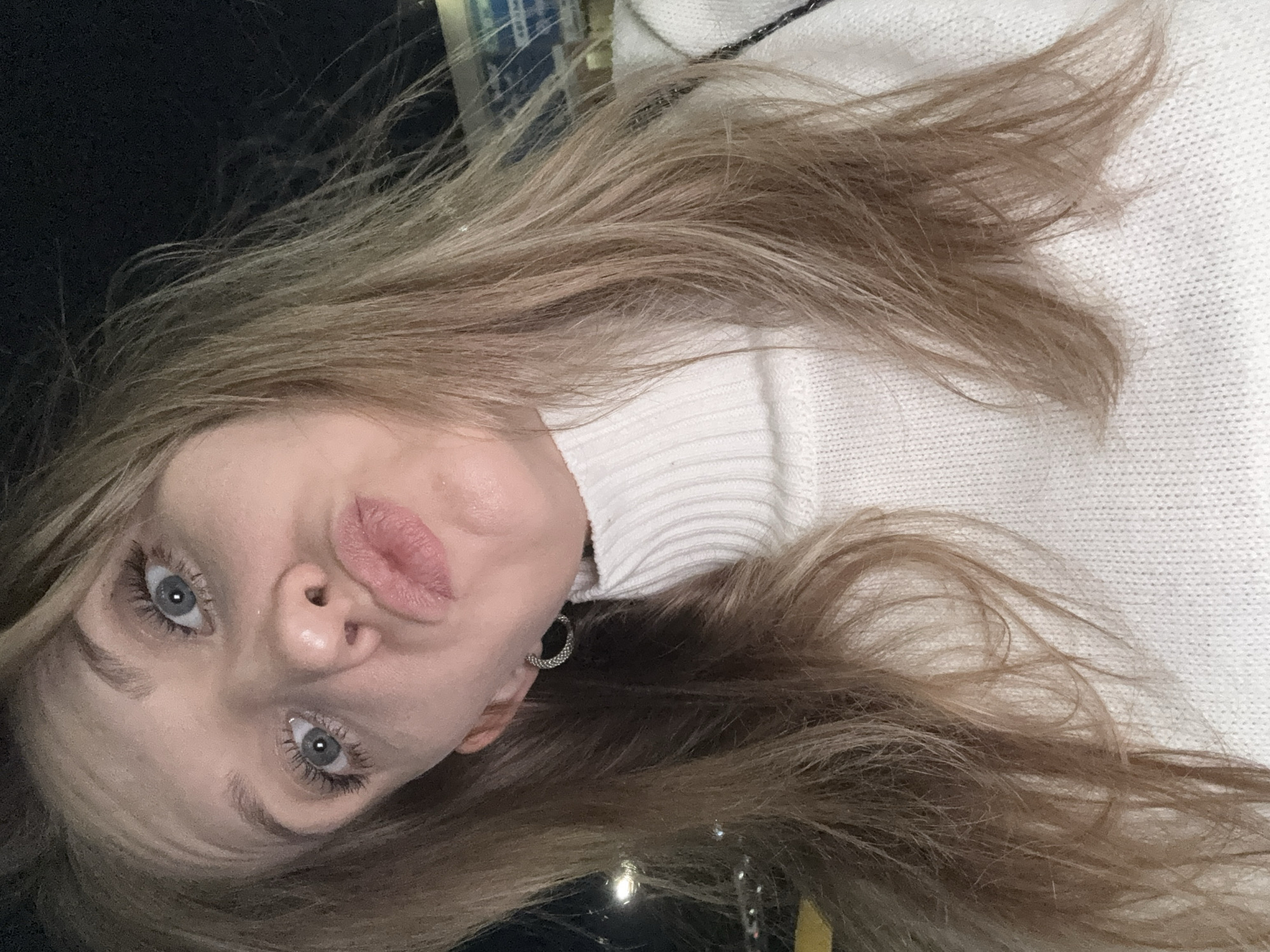 A woman with fair skin and gray eyes is facing the camera, her lips pursed in a kiss. She has long blonde hair and is wearing a white turtleneck sweater. A silver hoop earring is visible in her left ear. The background is dark, and there are a few blurred lights that may be reflections or streetlights. The location is not identifiable as Izhevsk, Russia, based on the image.