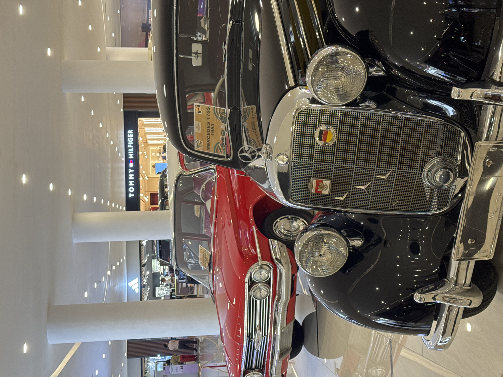 The image shows two classic cars, a black Mercedes-Benz 170S from 1953 and a red car of unknown make, parked in what appears to be a shopping mall. In the background, there is a Tommy Hilfiger store visible. The location is most likely in Indonesia, based on a sticker on the grill of the black car. The image focuses mainly on the front parts of the vehicles, especially the headlights and grills, while the rest of the environment appears to be a typical shopping mall setting with white columns and multiple shops. There are people visible in the background, suggesting the cars are on display.