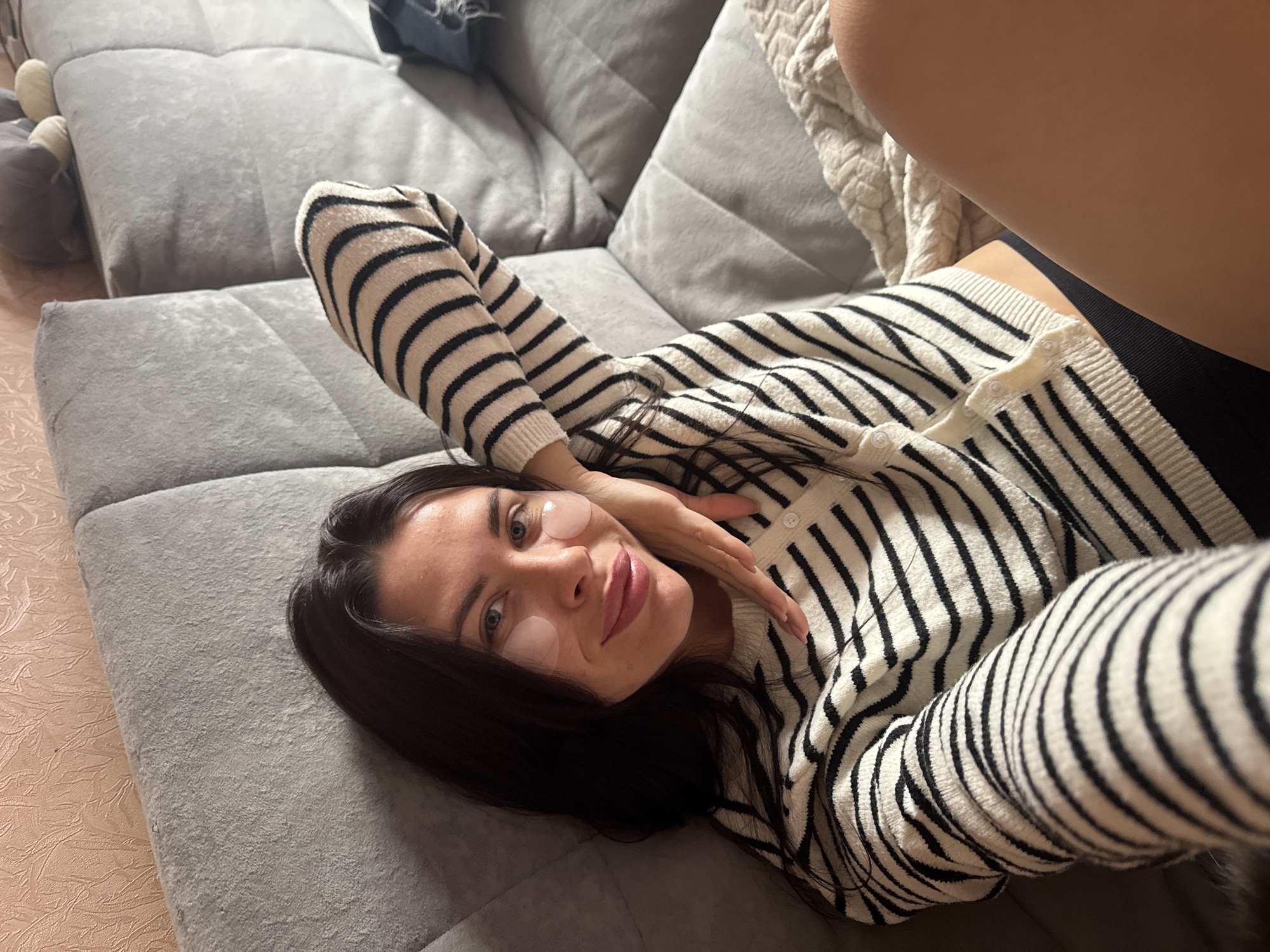 A woman with dark hair and eye patches is lying on a sofa. She is wearing a black and white striped sweater and black underwear. The sofa is gray and has a textured back. The wall behind the sofa is peach-colored and textured. There is a blanket on the sofa to the right of the woman. The lighting is dim. The image appears to have been taken indoors. The woman is the sole subject of the image. The image is a selfie. The woman is looking at the camera and smiling. Her left arm is resting on her head. The image is cropped to show her from the chest up. There are no other people or objects in the image that are clearly identifiable. The location in the city of Chaikovsky, Russia is not determinable from the image.