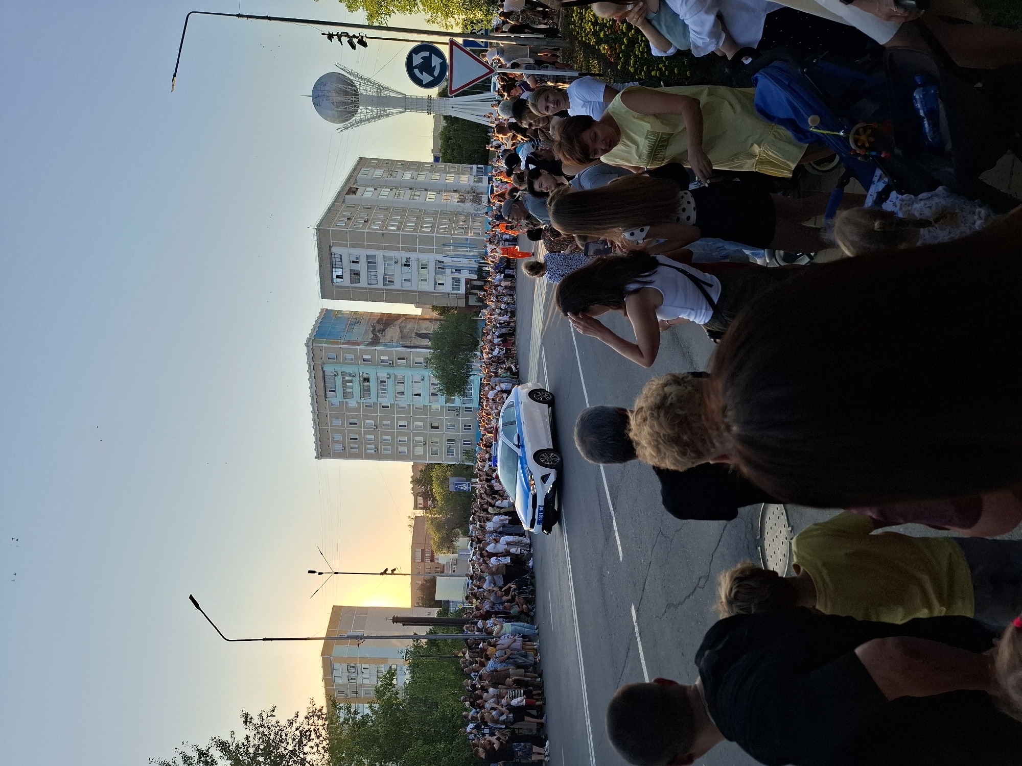 In an urban environment at dusk, a crowd of spectators lines a street, observing a procession. A white and blue police car, with lights illuminated, is moving down the road. On the left, apartment buildings and trees form the backdrop. On the right, a distinctive tall, spherical monument is visible, along with traffic signs. The sky is clear and light blue, indicating the setting sun is low on the horizon. People of various ages are gathered, some holding phones, watching the event unfold. The scene appears to be a public gathering or celebration in the city of Unknown.