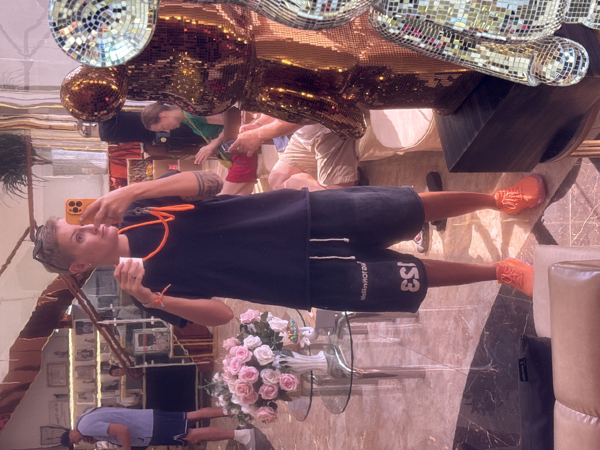 A person with short blonde hair is taking a selfie in a room with mirrored surfaces and reflective decorations. They are wearing a dark t-shirt and dark shorts, with orange sneakers. They are holding a white cup in their left hand and an orange cell phone in their right hand. An orange lanyard is around their neck. In the background, a man in a blue shirt and shorts is visible, as are several framed pictures on a wall. There is a vase of pink and white roses on a glass table. To the right, a large, bear-shaped sculpture covered in mirrored tiles is partially visible. The scene appears to be an indoor public space, possibly a lounge or lobby, in Nha Trang, Vietnam. The lighting is warm and reflective, creating a bright and somewhat dazzling atmosphere.
