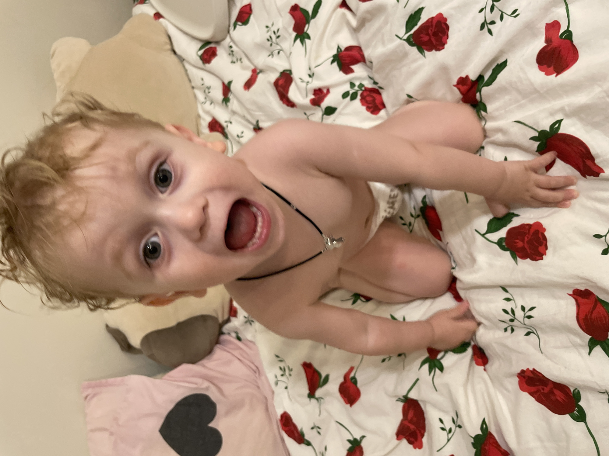 The image captures a lively moment with a young child, likely a toddler, positioned on their knees on a bed. The child, with light-colored, slightly curly hair, is looking directly at the camera with wide, expressive gray-blue eyes and an open mouth, suggesting they are mid-laugh, shout, or playful scream. Their face conveys strong excitement or surprise, with their tongue visible inside their mouth and a few bottom teeth showing.
The child appears to be wearing only a light-colored diaper or similar bottom wear, and a thin black cord necklace with a small, metallic, silver-toned charm around their neck. Their hands are resting on the patterned bedding.
The child is situated on white bedding adorned with a vibrant and detailed pattern of red roses and green leaves. To the upper left of the child's head, a beige, plush stuffed animal is partially visible, appearing to be its head and upper body. Further to the left and slightly below, a soft pink pillow with a large, solid black heart design is also seen. The background beyond these items appears to be a plain, light-colored wall.
The photograph is taken from an overhead perspective, looking down at the child, and is slightly tilted, giving a dynamic and candid feel to the snapshot. The indoor lighting is soft but sufficient, highlighting the child's features and the vivid colors of the bedding. The overall impression is one of candid joy, energetic engagement, and a moment of playful interaction captured from a personal memory collection.