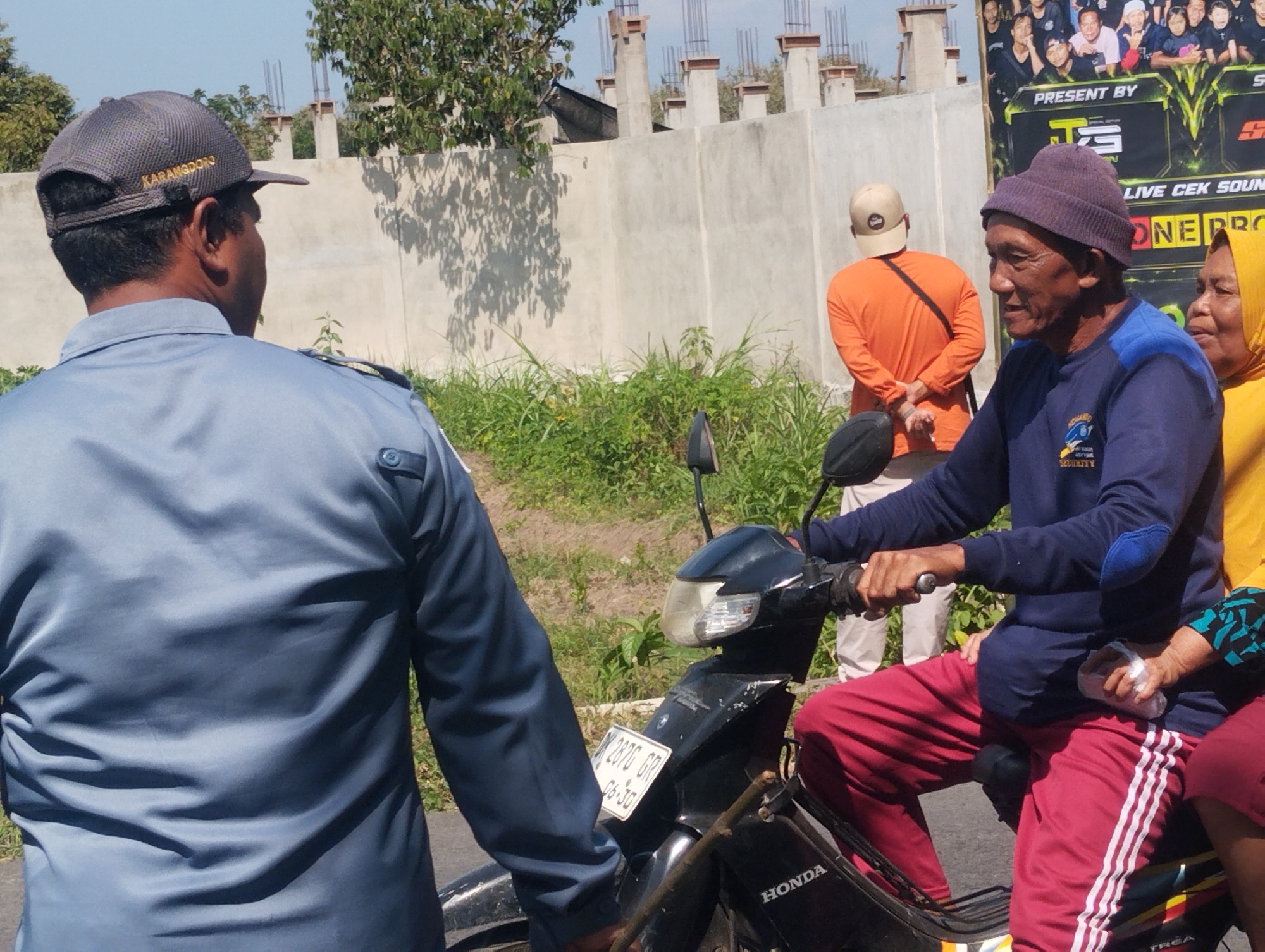 In the city of Karangdoro, Indonesia, on a sunny day, a man in a blue uniform and a grey cap is seen from the back, facing a motorcycle. He appears to be interacting with the individuals on the motorcycle. The man driving the motorcycle wears a purple beanie and a navy blue long-sleeved shirt with blue accents on the elbows. He is also wearing red track pants with white stripes. Seated behind him is a woman wearing a yellow headscarf. The motorcycle is black with a visible license plate that reads "W 2876 GR" and "06-30". In the background, a light grey wall with concrete pillars under construction runs horizontally. Lush green grass and foliage surround the area. A large banner with multiple faces and text, including "PRESENT BY TS" and "LIVE CEK SOUND", is visible on the right side of the image. Another man in an orange long-sleeved shirt and a beige baseball cap stands near the wall, facing away from the camera. The overall scene suggests a public gathering or event in an outdoor setting.