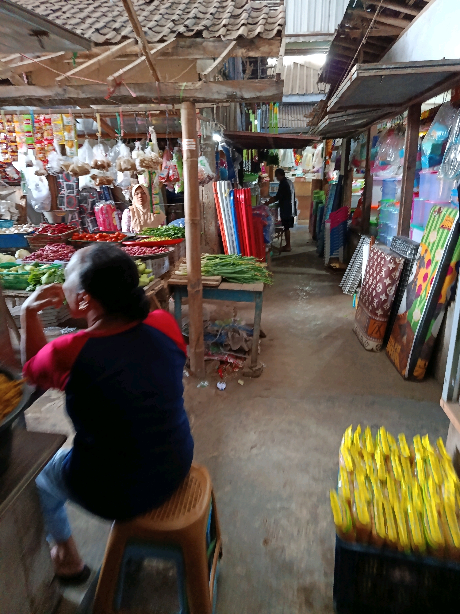 The image depicts a market scene in Mlawang, Indonesia. In the foreground, a person is seated on a plastic stool, facing away from the camera, with their back towards a stall. The person is wearing a dark blue t-shirt with red sleeves and light blue jeans. To the left of the person, there is a bowl containing yellow food items.
Behind the seated person, a market stall is visible, filled with various goods. A woman wearing a light-colored hijab and a patterned shirt is standing behind a display of vegetables and packaged goods. The stall is covered by a corrugated roof supported by wooden beams.
Further into the market, other stalls are visible, displaying a variety of items including colored plastic containers, rolled mats or fabrics, and what appear to be bottled drinks. Another person, dressed in dark clothing, is seen in the mid-ground, seemingly attending to a stall. The market floor is made of concrete. The overall environment suggests an open-air market with multiple vendors.