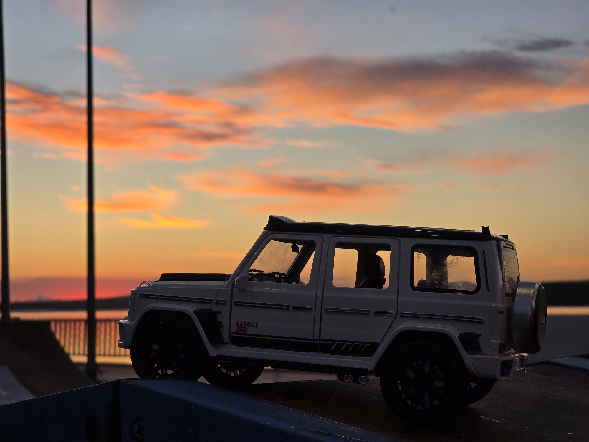 A white toy Mercedes-Benz G-Class, identifiable by the "BABOSI V12" decal on its side, is positioned on a ramp at a skatepark during a sunset. The sky is painted with vibrant orange and pink clouds against a pale blue backdrop, suggesting a warm evening. In the background, a body of water and a railing are visible. The toy vehicle's details, including its spare tire and exhaust pipes, are clearly depicted. The lighting creates a silhouette effect on some elements of the scene. The image appears to be taken in Chaikovsky, Russia, a city known for its industrial and recreational facilities.