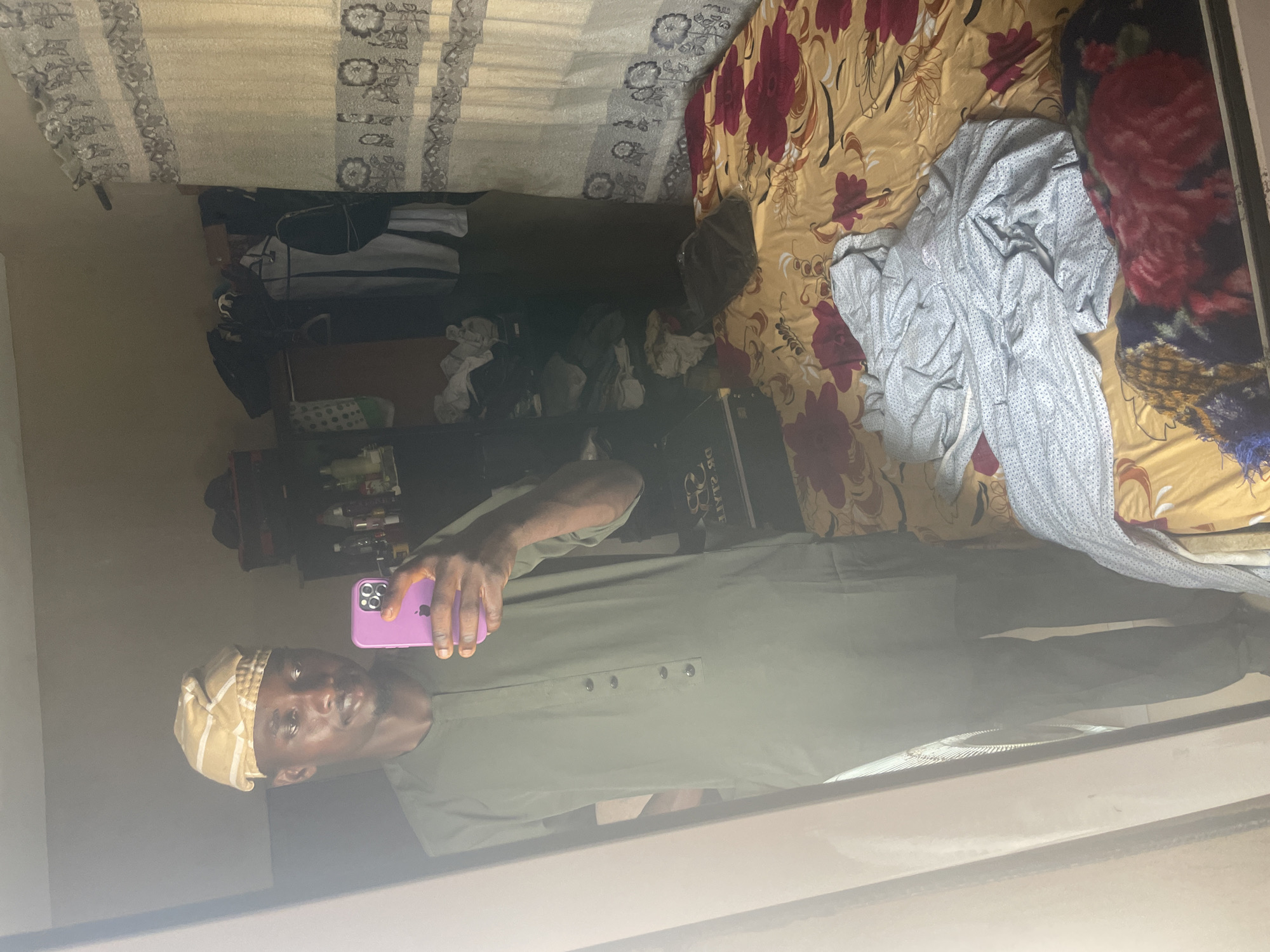 A man stands in front of a mirror, holding a smartphone and taking a selfie. He is wearing a light green garment with a head covering. The reflection shows a cluttered room with a bed, bedding, and other objects on shelves. The image was taken in an interior space in Badagry, Nigeria.