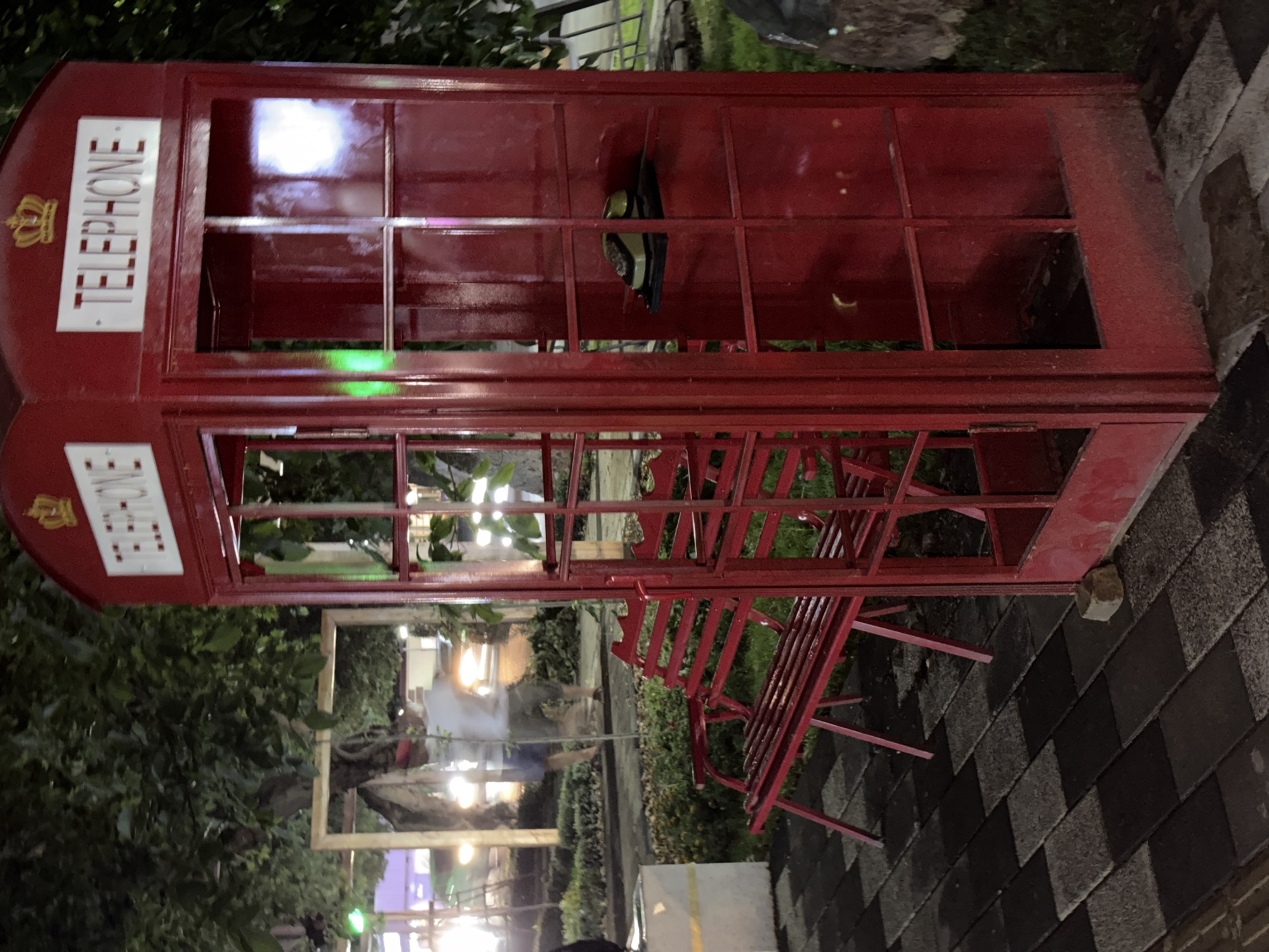 The media file shows a red British-style phone booth, oriented vertically, in an outdoor setting. The phone booth has glass panels and the word "TELEPHONE" in white lettering with a crown emblem above it on one side. Inside the booth, a red metal bench is visible, as well as some greenery. The booth is situated on a patterned tiled surface composed of dark grey and black squares. In the background, there are trees and bushes, with some lights visible, suggesting it might be evening or nighttime. The overall scene appears to be an outdoor public space, possibly a park or a decorative area in Chirchik, Uzbekistan. There are no people clearly visible, but the scene suggests a place that might be visited or used. The phone booth itself is a prominent object, while the surrounding environment includes natural elements like trees and man-made elements like the tiles and the bench. The lighting is somewhat dim, with reflections on the glass panels of the phone booth, hinting at artificial light sources nearby.