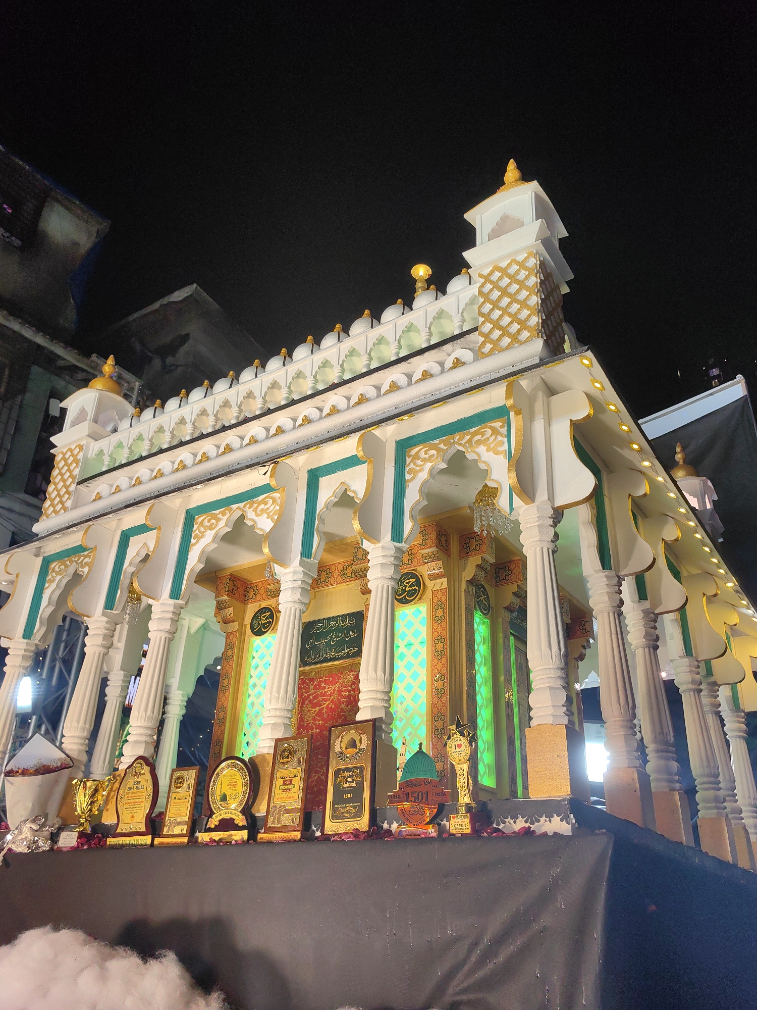 The image shows a brightly lit, ornate structure, likely a temporary display or shrine, at night. The structure is predominantly white with gold and turquoise decorative accents, featuring arched openings and columns. It is adorned with religious calligraphy and illuminated with green lights from within. In front of the structure, a display of trophies and awards is arranged on a black surface, some of which are illuminated by spotlights. The background reveals a dark sky, with some indistinct structures visible to the left, suggesting an urban setting. The presence of the trophies and the celebratory decorations indicates an event or commemoration. The image was taken from a low angle, looking upwards at the structure. The location is identified as Mumbai, India.