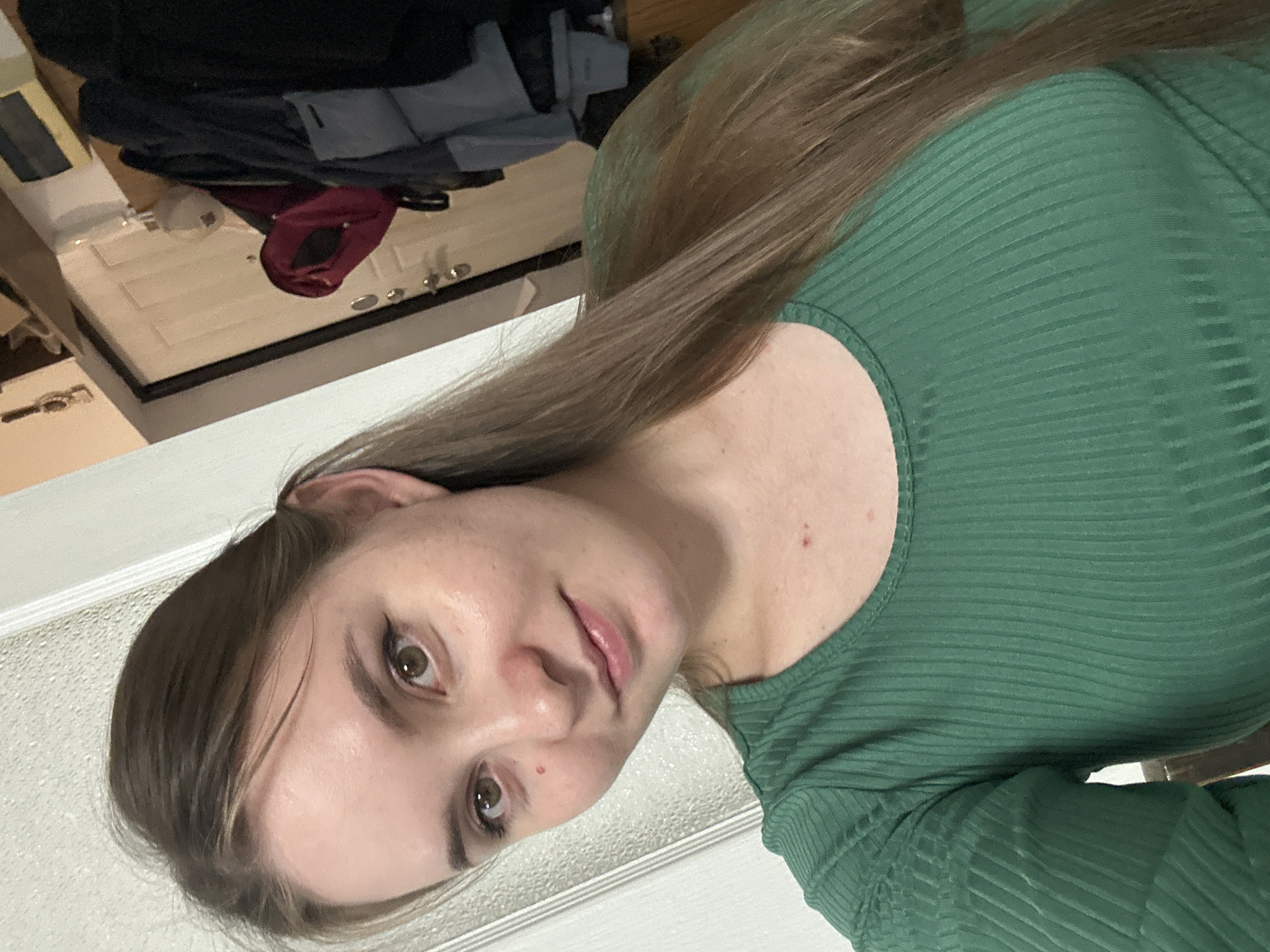 A young woman with light skin, brown hair, and brown eyes is taking a selfie indoors. She is wearing a green, ribbed, long-sleeved shirt. Her hair is pulled back into a ponytail. She has eyeliner on and a mole on her cheek. In the background, there is a white door with a dark handle, a shelf with various items, and some coats hanging on a rack. The setting appears to be a home. The specific location in Noviy, Russia is not discernible from the image.