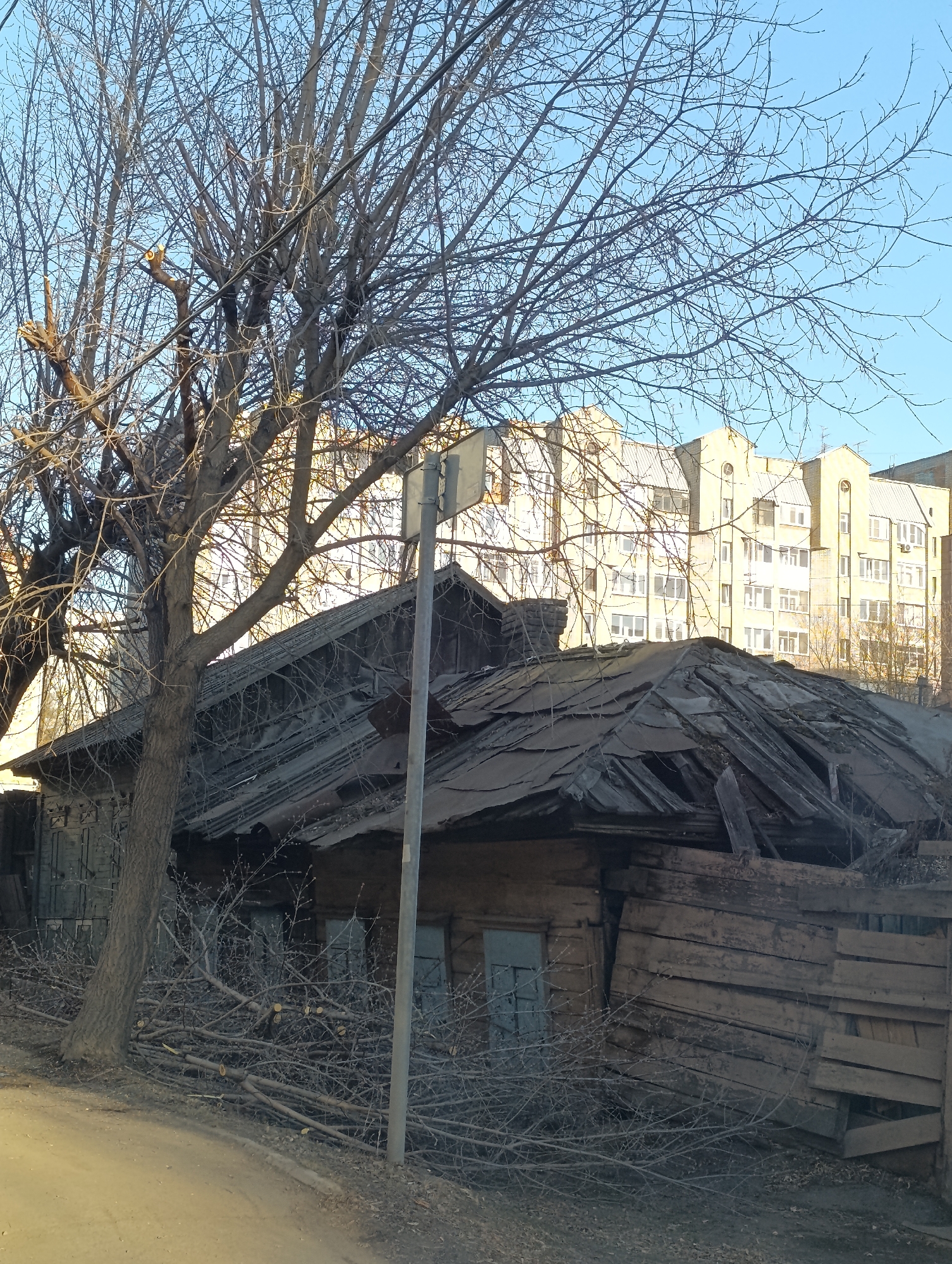 The image depicts a dilapidated wooden house in Krasnoyarsk, Russia, against a backdrop of a modern apartment building. The house shows significant disrepair with a collapsing roof, weathered wooden siding, and boarded-up windows. A bare tree stands in front of the house, and cut branches are piled on the ground near the base of the tree. A utility pole is positioned close to the house. The sky is clear and blue. A paved road borders the scene's left side.
