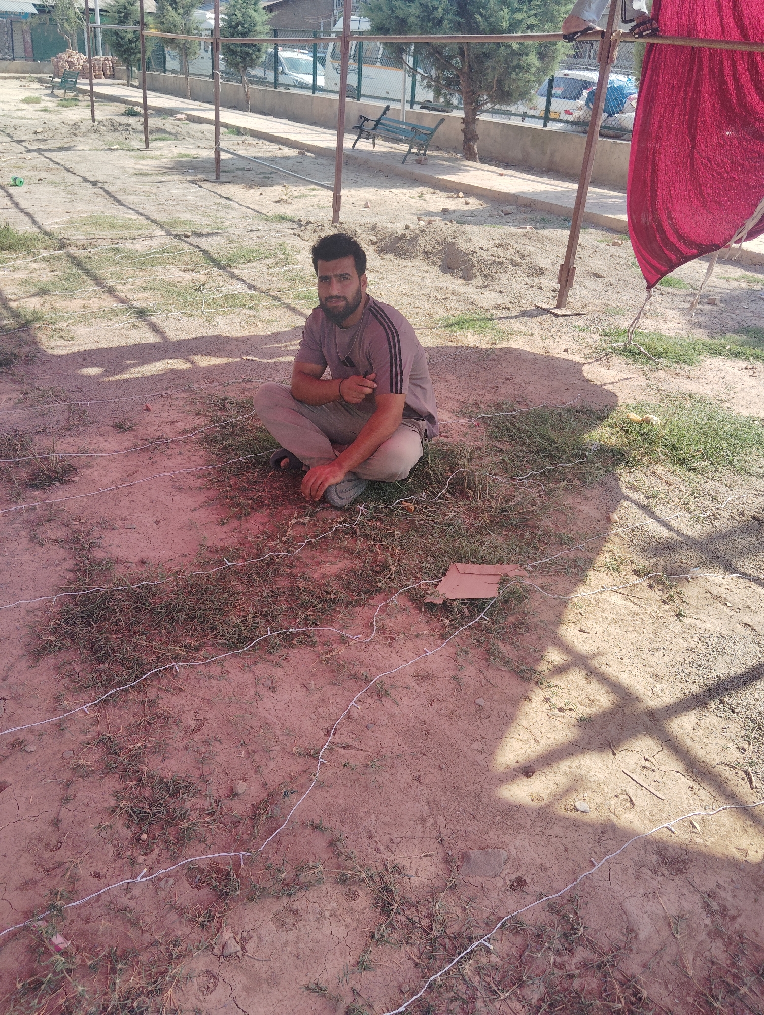 A young man with dark hair and a beard sits cross-legged on the ground in a fenced outdoor area. He wears a grey t-shirt with black stripes and light-colored pants. The ground is a mix of dry dirt and sparse green grass, with several thin white lines laid out in a grid pattern on the surface. In the background, there is a chain-link fence, some trees, and what appears to be a stone wall. Further back, a white vehicle and more trees are visible. A large piece of red fabric hangs from a pole on the right side of the frame. The scene suggests an outdoor recreational or event space, possibly within the city of Srinagar, India, given the vegetation and architectural elements.