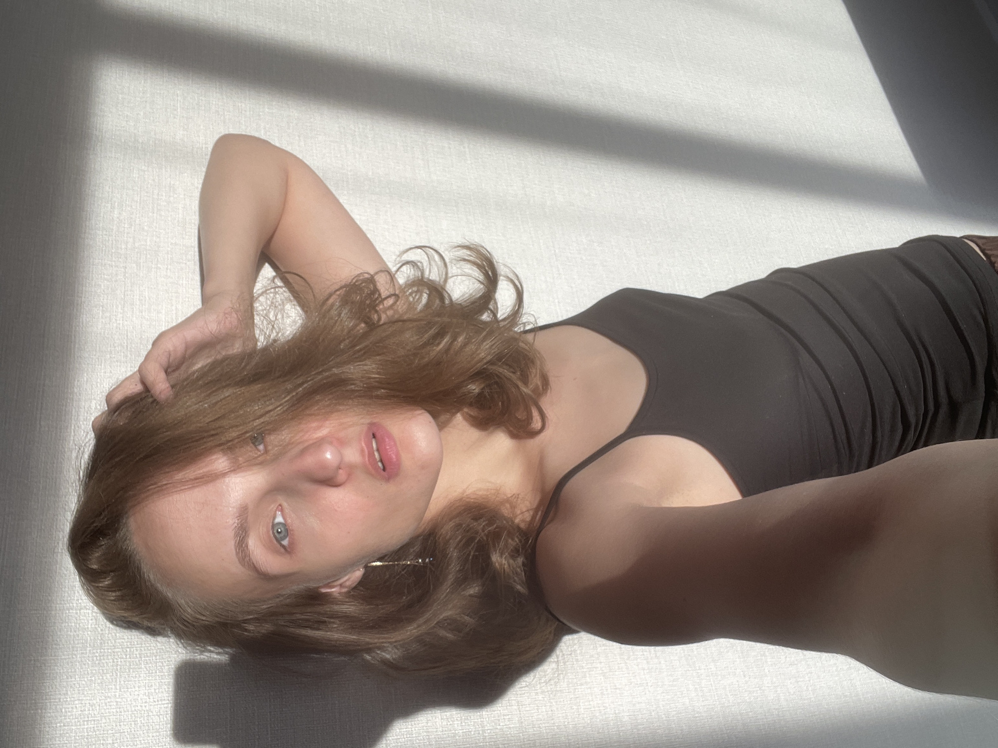 This media file captures a close-up, overhead shot of a young woman lying on her back. She has shoulder-length, wavy brown hair, fair skin, and light blue eyes. She is wearing a black tank top. Her left arm is bent at the elbow, with her hand resting on the back of her head, and her right arm is extended outwards, out of frame, suggesting she may be taking the photo herself. The woman's head is tilted back, and her lips are slightly parted, with her mouth open, revealing her teeth. She has a delicate, almost wistful expression.
The scene is illuminated by strong, directional sunlight, casting sharp shadows across the white textured surface on which she is lying. The shadows suggest the presence of an object, possibly a window frame or blinds, indicating an indoor setting bathed in natural light. The light creates a dramatic interplay of highlights and shadows across her face and body, enhancing the mood of the image. The overall atmosphere is serene and intimate, with a sense of quiet contemplation. The time of day appears to be late morning or early afternoon, given the angle and intensity of the sunlight. There is no visible text in the image. The location context provided is Подшивалово, Russia.