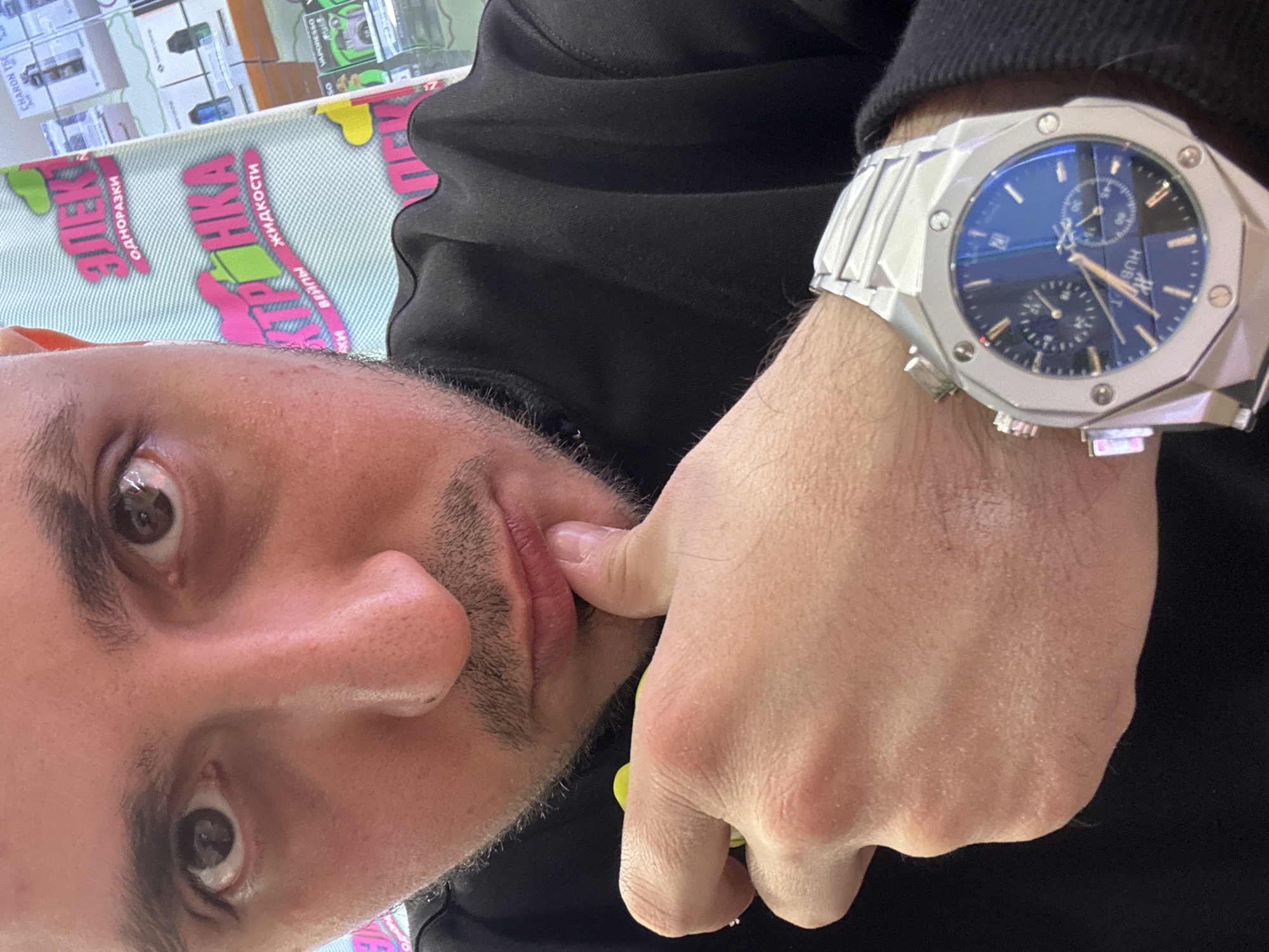 A man is posing for a selfie, his face close to the camera. He has light skin, dark hair, and a mustache. He is wearing a black shirt. His left hand is raised to his face, and he is touching his lip with his index finger. On his left wrist, he is wearing a silver Hublot watch with a blue face. The background is blurry but appears to be a retail setting. A sign in Russian is visible, suggesting the location is likely in Russia, possibly Saint Petersburg given the signage.
