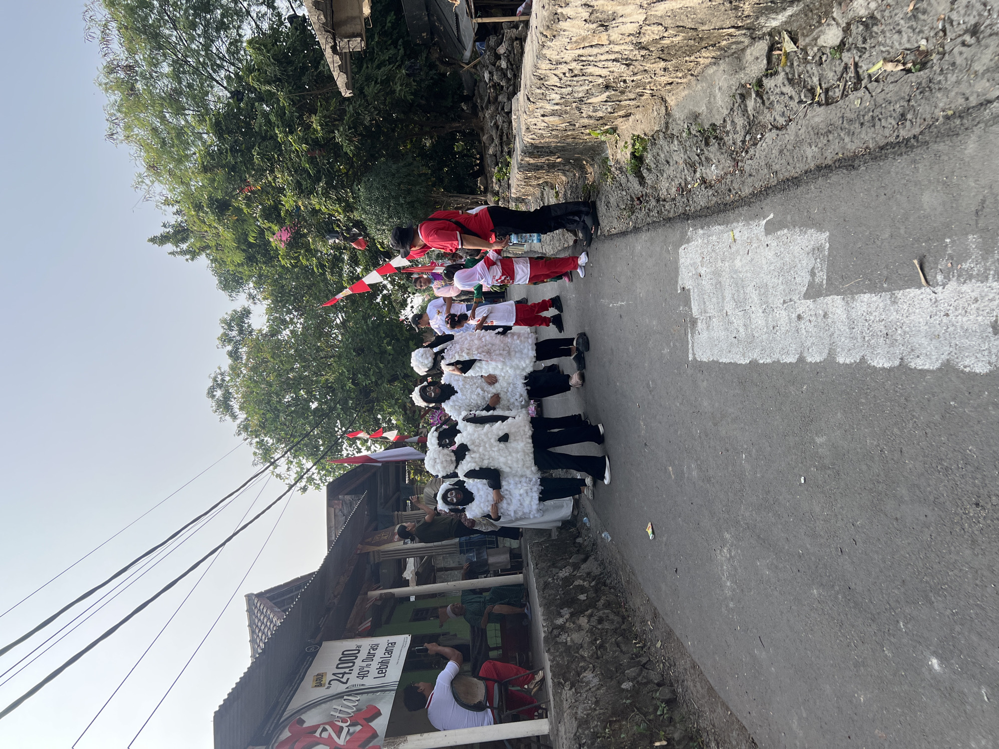 In Klapanunggal, Indonesia, a street scene captures a group of individuals in sheep costumes participating in a procession. The individuals are wearing white, fluffy costumes with matching headwear, their faces painted black. They are walking on an asphalt road lined with stone walls and vegetation. To the left, a building with a sign that reads "BARU 24.000" is visible. Several people are watching the procession from the side, some holding cameras. Indonesian flags are displayed on poles in the background. The sky is overcast, and the lighting suggests it is daytime.