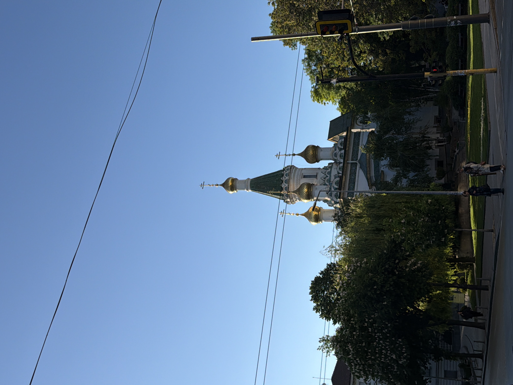 The image depicts the Church of St. Nicholas the Miracle-Maker in Sofia, Bulgaria. The church has distinctive gold domes and green tiled roofs. Power lines are visible against the clear blue sky. A traffic light pole stands nearby. Trees and a patch of grass line the street. Two people are walking along the sidewalk in the background.