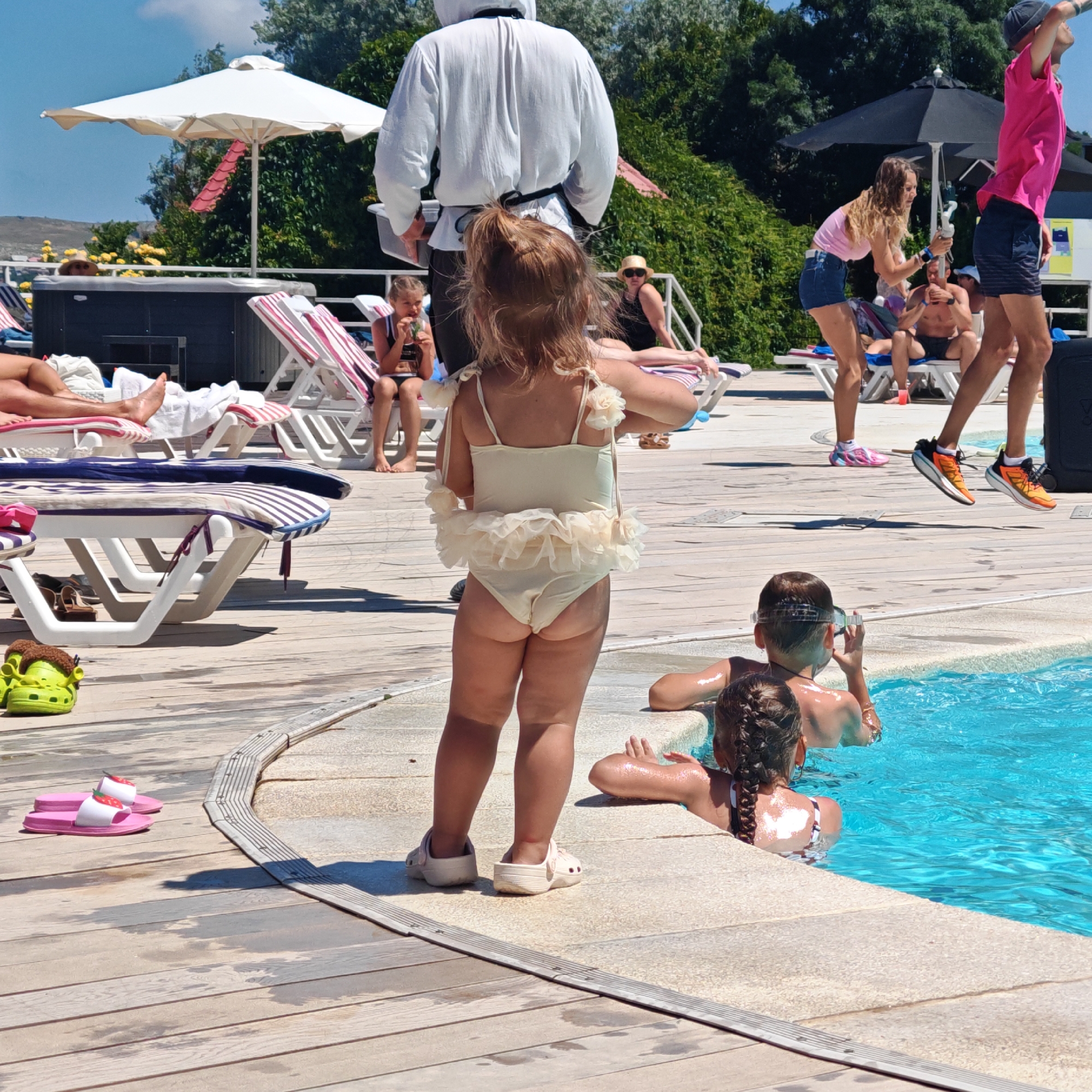 This image depicts a sunny outdoor scene, likely at a pool or resort, with several people present. In the foreground, a young girl in a light-colored swimsuit with ruffles is standing, facing away from the camera. To her left, a pair of pink slides with strawberry designs rests on the wooden deck. Further back, other individuals are visible, some relaxing on lounge chairs, and a few are in the bright blue pool. The background includes lush green trees, a white umbrella, and a black umbrella. The overall environment suggests a recreational setting.
