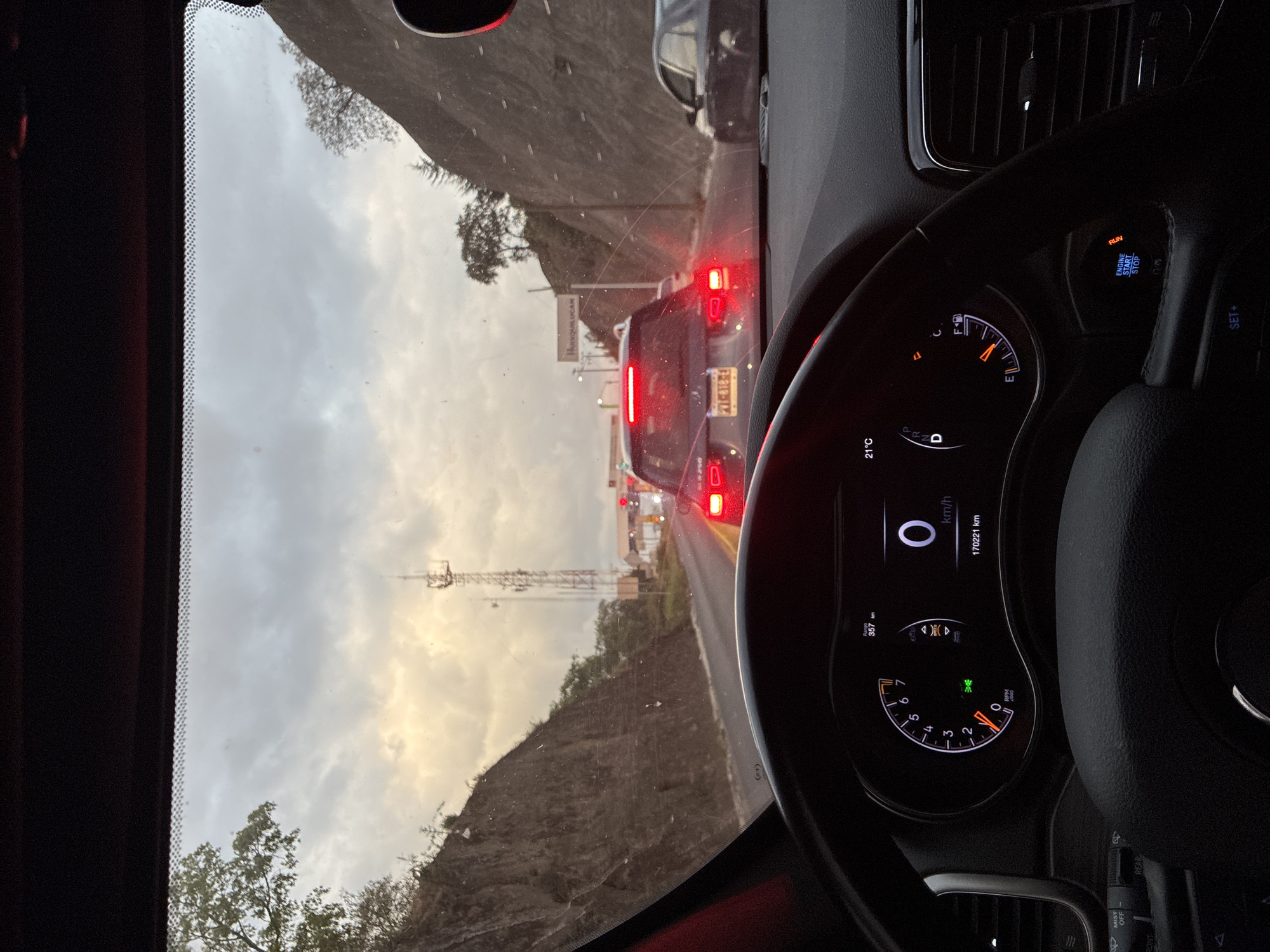 The image shows a view from inside a car, likely a Jeep, stuck in traffic in Huixquilucan de Degollado, Mexico. The dashboard indicates the car is stationary (0 km/h) and the engine is running ("RUN" is illuminated). The odometer reads 170221 km. The fuel gauge is near empty. The ambient temperature is 21°C. Outside, the sky is overcast. Visible through the windshield are other vehicles in the traffic jam, including a dark-colored vehicle with the license plate PVT-818-E1. A sign with "Republican" is visible. The landscape includes a hillside and communication towers.