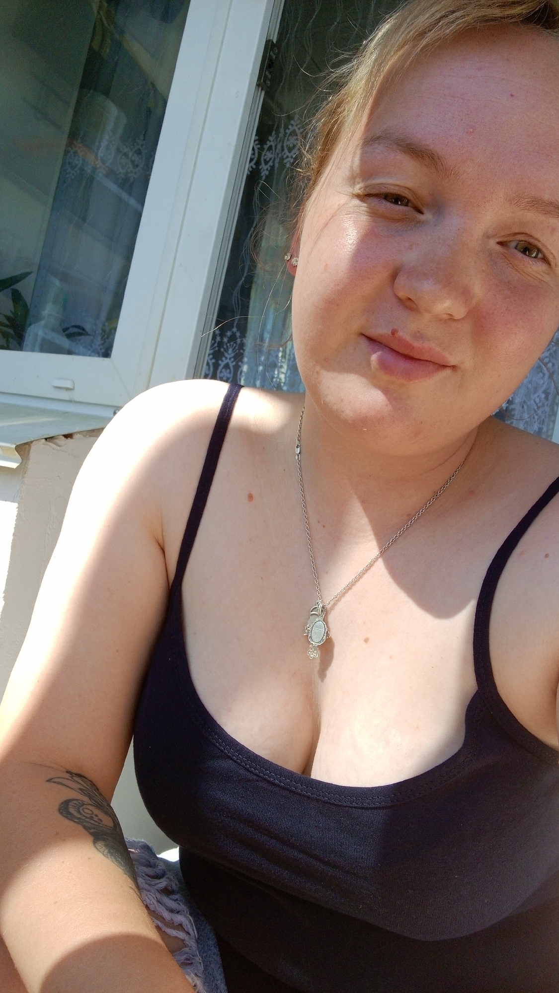 A selfie taken in Уральское, Russia shows a fair-skinned woman with light blonde hair pulled back, wearing a black tank top and a silver necklace with a pendant. She is positioned outdoors, likely on a porch or balcony, as a white window frame with a lace curtain is visible in the background. The lighting suggests it is daytime, possibly sunny. The woman has a slight smile and is looking directly at the camera. A tattoo is visible on her upper left arm.