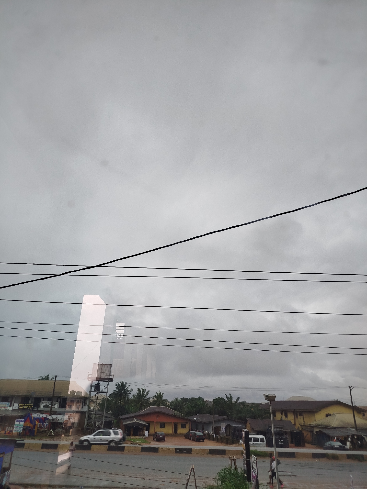 The image captures a street scene in Uselu, Nigeria, under a cloudy sky. A main road runs horizontally across the frame, with buildings and vegetation lining its side. Several cars are visible, including a silver SUV and a white van. Some people are walking on the sidewalk and street. Overhead, power lines crisscross the sky, and a reflection of a tall, modern building is superimposed on the scene. The architecture of the buildings appears to be a mix of residential and commercial structures, with corrugated metal roofs being a prominent feature. The overall impression is that of a typical urban environment in Nigeria, with evidence of infrastructure such as power lines and roads.