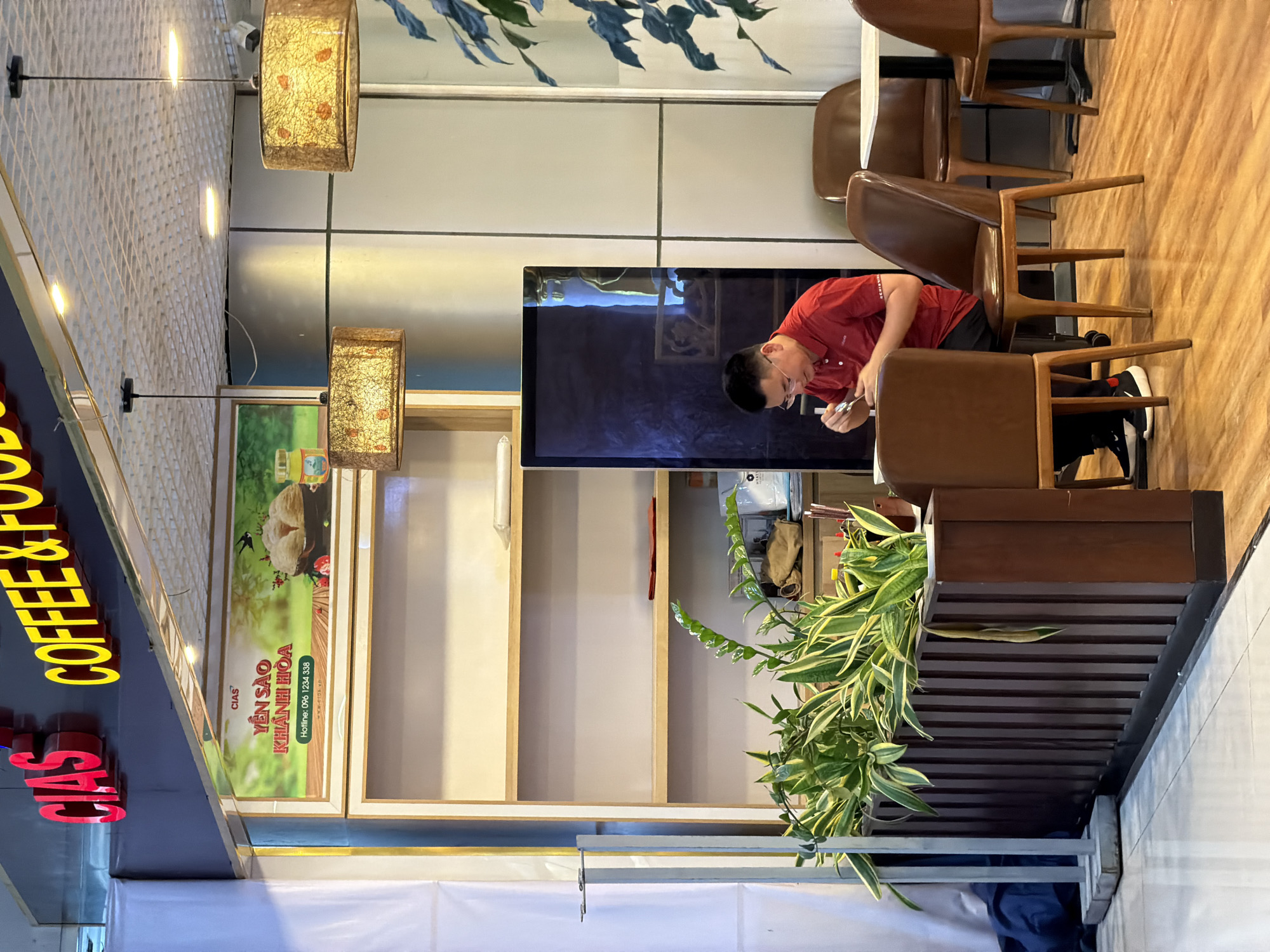 A man wearing a red polo shirt is seated at a small round table in a cafe, engaged in eating. He is positioned in the foreground, right of center, with his back slightly angled towards the viewer. His head is lowered as he uses cutlery. The cafe interior features wooden floors, and several chairs and tables are visible. A tall wooden partition with potted plants stands to the left of the man. Behind him, a wall with grid-like patterns and a vertical dark panel are present. Above, two decorative lamps with translucent shades hang from the ceiling. A sign reading "CIAS COFFEE & FOOD" is prominently displayed at the top of the frame. To the left of the man, a display area with shelves is visible, and a large advertisement for "YEN SAO KHANH HOA" is mounted above it. The overall ambiance suggests a casual dining environment.