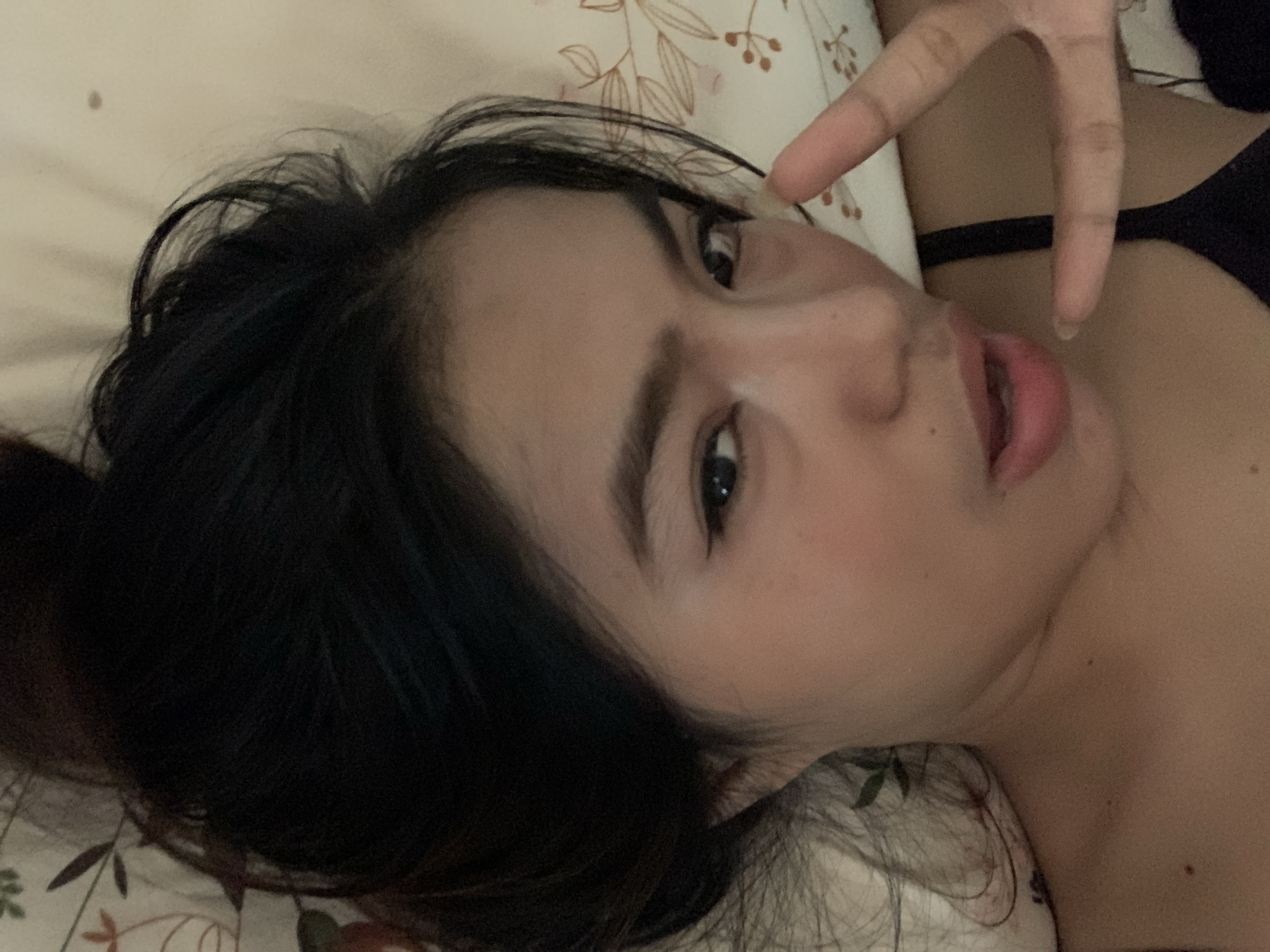 A close-up selfie shows a young person, likely female, lying down on a light-colored surface, possibly a bed, with their head tilted. The person has long, dark, slightly messy hair that frames their face and spreads across the pillow. They are looking directly into the camera with dark eyes, which have visible eyeliner. Their mouth is slightly open, revealing their tongue.
One hand is raised near their face, making a peace sign with the index and middle fingers extended. The skin tone appears fair, and a few small moles are visible on the chest and shoulder area. A black strap, indicative of a bra or camisole, is visible over their right shoulder. The background consists of a pillow or sheet with a subtle, light-colored floral pattern featuring brown and green leaf motifs. The lighting suggests an indoor setting, though the exact time of day (e.g., day or night) is not explicitly clear. The overall expression conveys a casual and possibly playful mood. There is no visible text in the image.