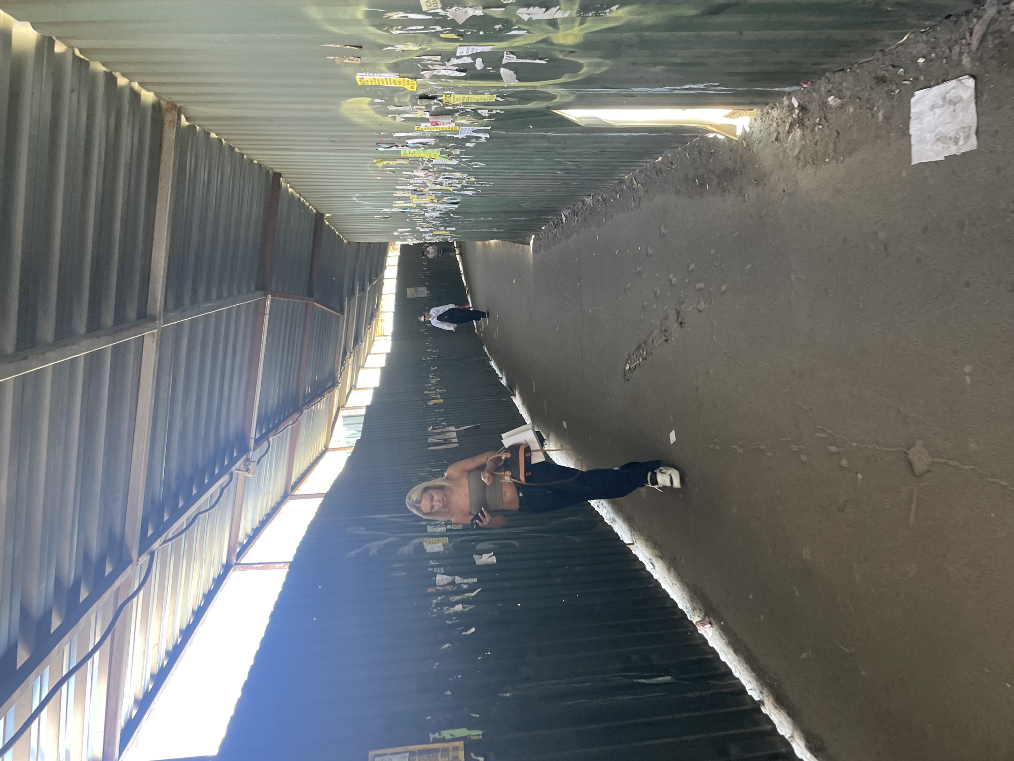 The image shows a narrow, covered walkway with corrugated metal walls and a corrugated metal roof. The walkway appears to be outdoors and is lit by sunlight filtering through gaps in the roof. The walls are covered in graffiti and posters. In the foreground, a young woman with blonde hair is walking away from the camera, looking at her phone and carrying a small handbag. She is wearing a strapless top, dark pants, and white sneakers. Further down the walkway, another person is visible walking away from the camera. The ground is paved and appears somewhat worn. The overall impression is of a somewhat utilitarian and possibly neglected urban space. The location is identified as Kyiv, Ukraine.