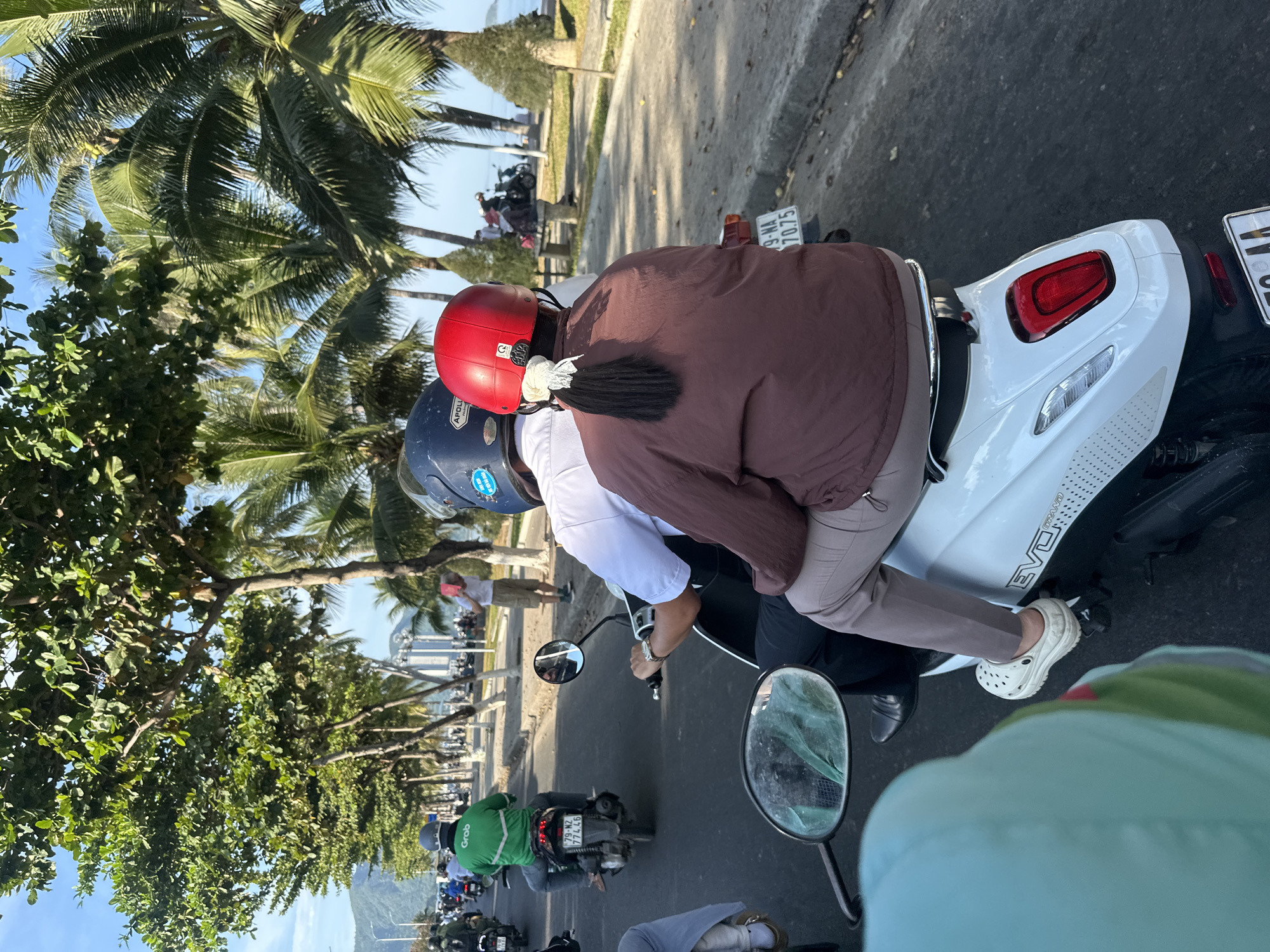 The image depicts a scene on a street in Nha Trang, Vietnam, characterized by a sunny day with a clear blue sky. The foreground shows the rear of a white scooter, with a person wearing a brown jacket and tan pants as the passenger. The passenger has a red helmet and their hair is tied in a ponytail with a white scrunchie. The scooter's license plate is partially visible.
In the middle ground, the scooter is being ridden by a person wearing a white shirt and a dark helmet. They are looking forward, presumably driving. The road is asphalt, with a slight incline. To the right of the road, there's a paved sidewalk with trees and palm trees lining it, indicating a coastal area or park. Some people are visible on the sidewalk, one of whom is standing and holding a pink object to their face.
Further down the road, there's a line of other scooters and people, suggesting moderate traffic. One scooter rider in the distance is wearing a bright green jacket with the "Grab" logo on the back, a common ride-hailing service in Southeast Asia. The background shows buildings and a distant hillside, characteristic of Nha Trang's cityscape, with the ocean visible further in the distance. The overall impression is a typical street scene in a Vietnamese coastal city during the day.