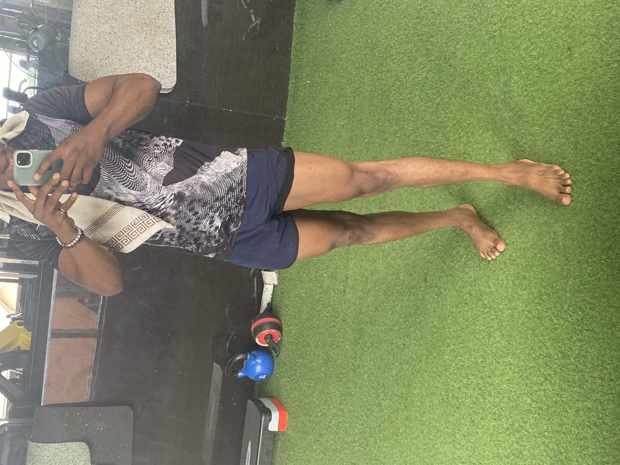 A person stands on artificial turf in what appears to be a gym. The person is wearing a dark t-shirt with a white graphic design, dark blue shorts with a black trim, and a light-colored towel draped over their shoulders. They are holding a smartphone in their right hand, taking a selfie. To the left of the person are a black aerobic step with red and white accents, and a blue kettlebell. To the right of the kettlebell is a red and black ab roller. The background consists of a black wall and some gym equipment, including a large fan on the right. There is no visual information in the image that could indicate the specific location in Podo, Nigeria.