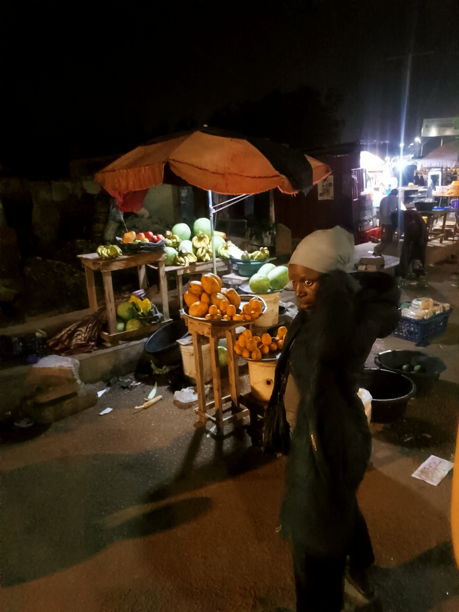 The image depicts an outdoor fruit market in Bukuru, Nigeria, at night. A woman wearing a gray head covering and a dark coat stands in the foreground, partially obscuring the view. Behind her, fruit is displayed on tables under an orange and black umbrella. Fruits include bananas, watermelons, mangoes, and tomatoes. Other market stalls are visible in the background, illuminated by electric lights. Debris litters the ground.