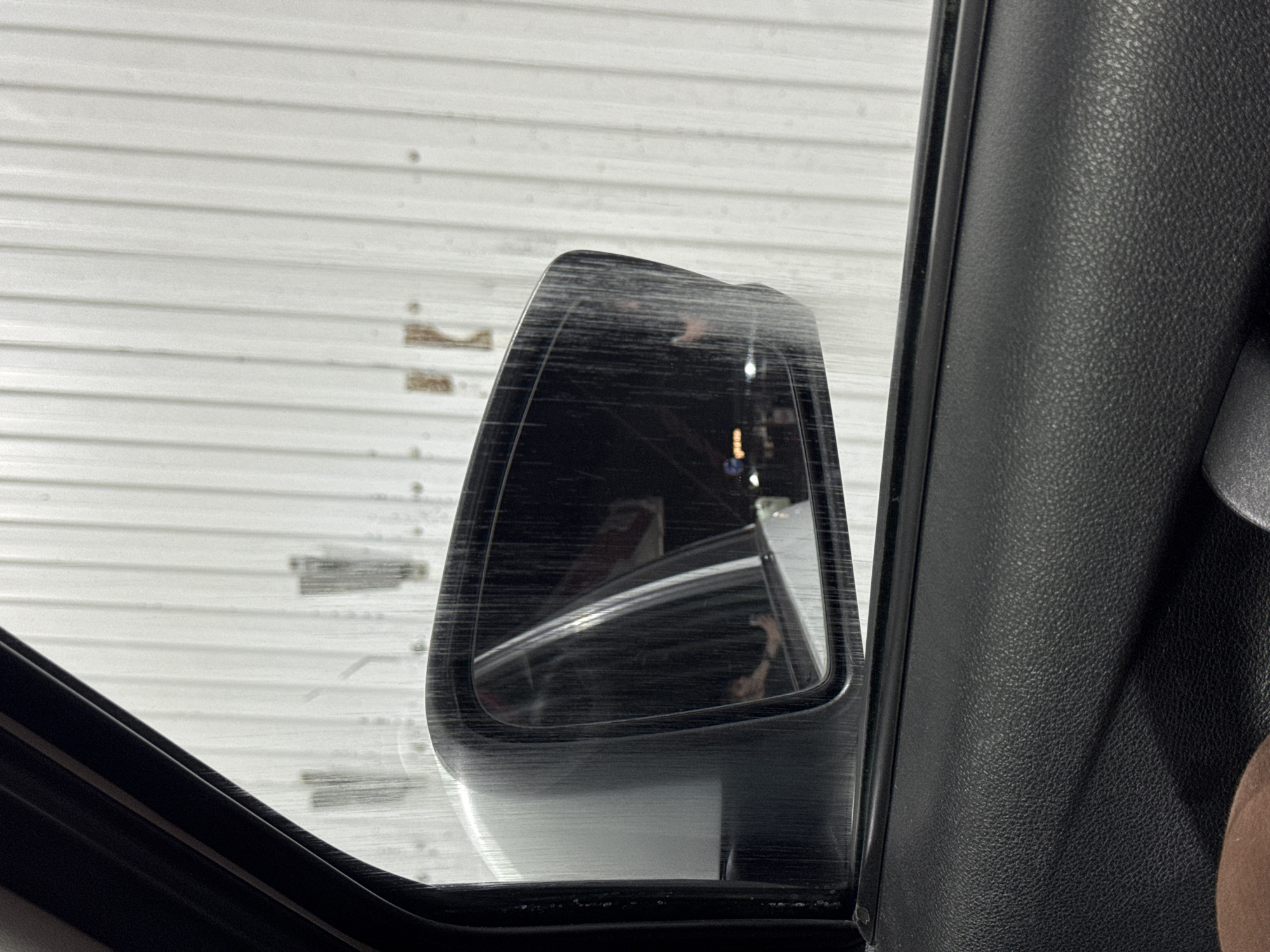 The image shows a view from inside a vehicle, looking out the driver's side window. A side-view mirror reflects the interior of the car, showing a person's hand and arm reaching towards the mirror. The background outside the car is a ribbed white metal wall with some faint marks on it. The car interior visible includes a black textured door panel and part of the window frame. The presence of what appears to be rain streaks on the window and mirror suggests it may be raining or the car is wet. The image context does not provide information about the specific location within Ilskhan-Yurt, Russia.