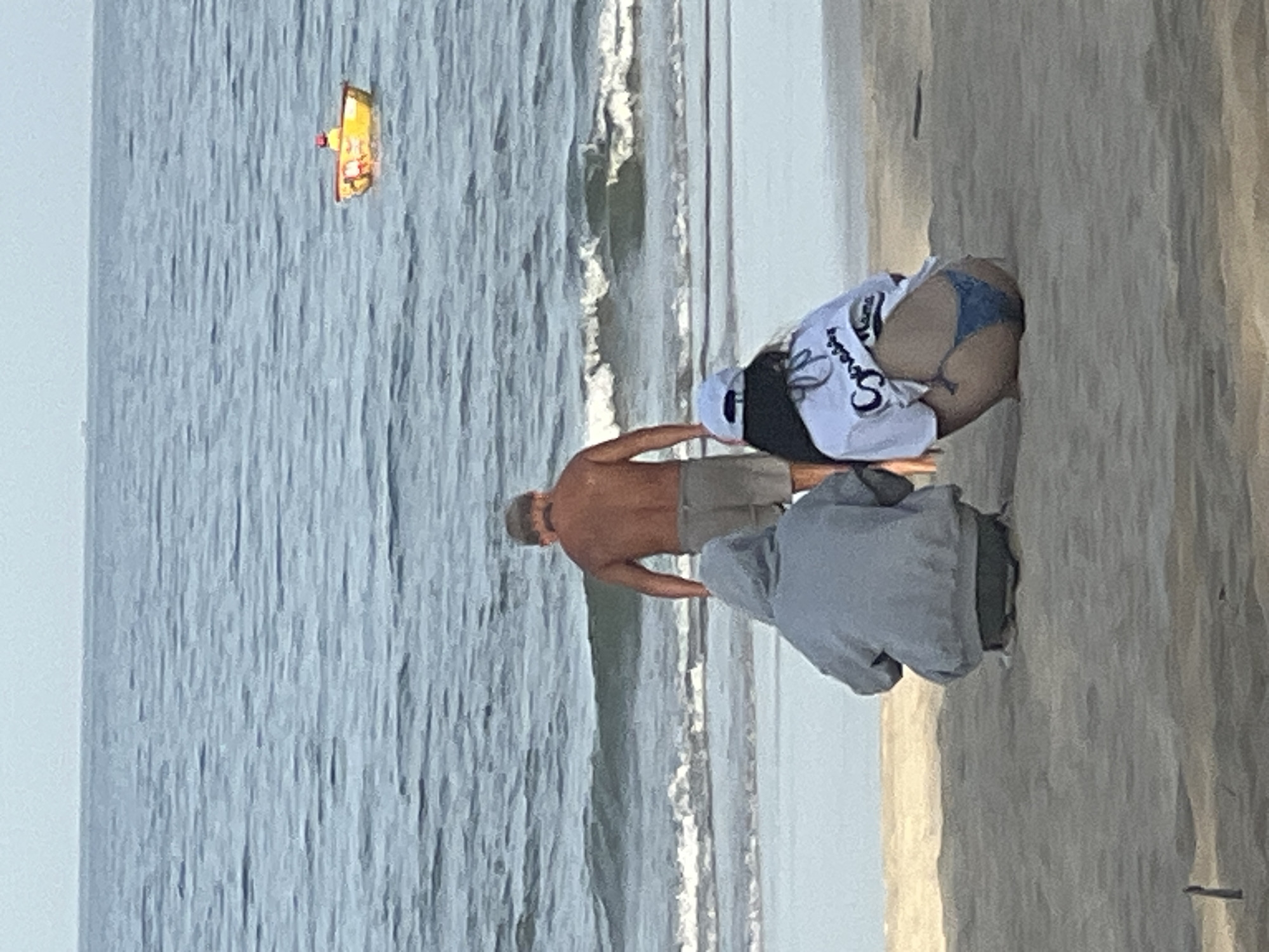 A medium shot captures a beach scene with three individuals near the water's edge on a sandy shore. The ocean, a hazy blue, stretches to the horizon under a pale sky. In the distance, a small, yellow raft with a figure is visible on the water.
In the foreground, two individuals are crouched on the sand, facing away from the camera towards the sea. The person on the left wears a gray hooded sweatshirt and dark pants. The person on the right, also facing away, wears a white t-shirt with dark lettering on the back and a light blue bikini bottom.
A man stands between them, facing the ocean. He has short hair and is wearing light-colored shorts. He is positioned closer to the water, with waves breaking behind him. The overall impression is a casual beach outing. The location is unspecified in the image.