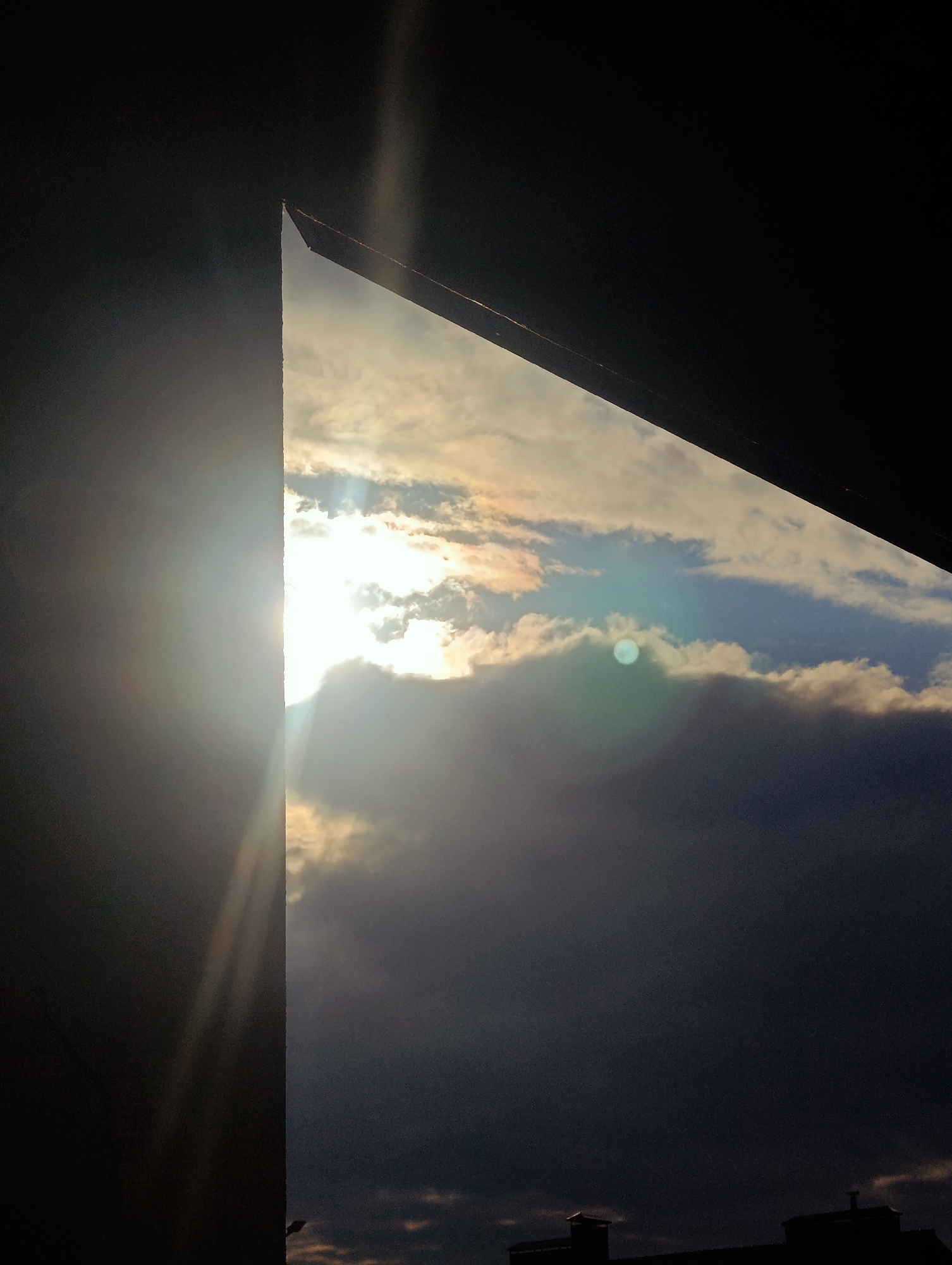 The image depicts a dramatic sky with the sun partially obscured by clouds, seen from a vantage point within a city. The sun's rays create a bright lens flare on the left side of the frame, casting beams of light through gaps in the clouds. The sky transitions from bright white and yellow near the sun to various shades of blue and gray, with darker, heavy clouds dominating the lower portion of the image. The silhouette of a building or structure with a roofline is visible at the very bottom of the frame, suggesting an urban environment. The scene is characterized by strong contrasts between light and shadow, likely captured during sunrise or sunset.