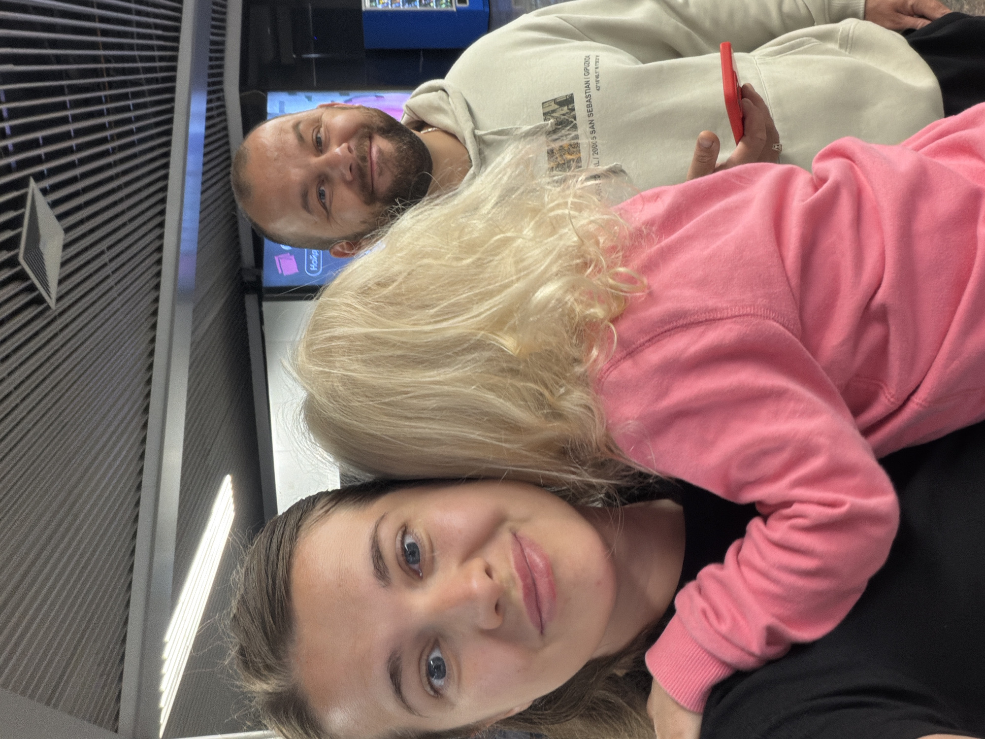 The image shows a family, a woman, a man, and a child, posing for a selfie indoors. The woman is in the foreground, looking directly at the camera with a slight smile. A blonde child, facing away from the camera, is embracing the woman. Behind them, a man with a beard is smiling and looking at the camera. The background suggests an indoor public space, possibly an airport or train station, with a ceiling featuring metallic ribbed panels and some lighting fixtures. A vending machine is visible to the right. The location is identified as Sochi, Russia.