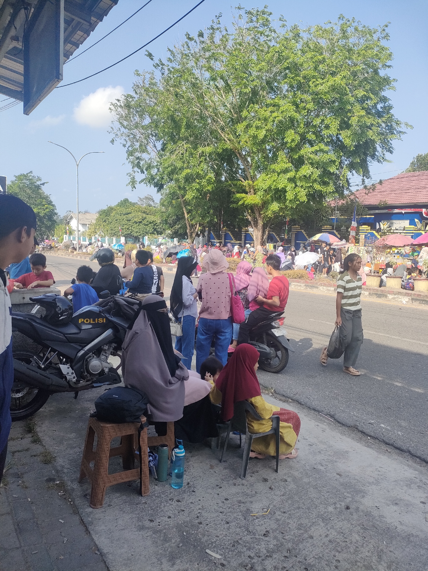 In Sungailiat, Indonesia, on a sunny day, a crowd gathers along a street. The scene is characterized by a mix of individuals, some standing and others seated on chairs and stools, indicating a temporary waiting area or gathering point. A prominent black motorcycle with "POLISI" visible on its side is parked, suggesting a law enforcement presence or activity. Several people are seen near the motorcycle, including children and adults.
The environment appears to be a roadside with trees providing shade. In the background, buildings with red roofs are visible, along with more trees and a clear blue sky. The ground is paved, with a grassy area to the side. Many people are dressed in casual clothing, and some women are wearing hijabs. The presence of umbrellas and parked motorcycles further suggests a local event or gathering. The overall atmosphere seems to be one of a public space being utilized by a community.