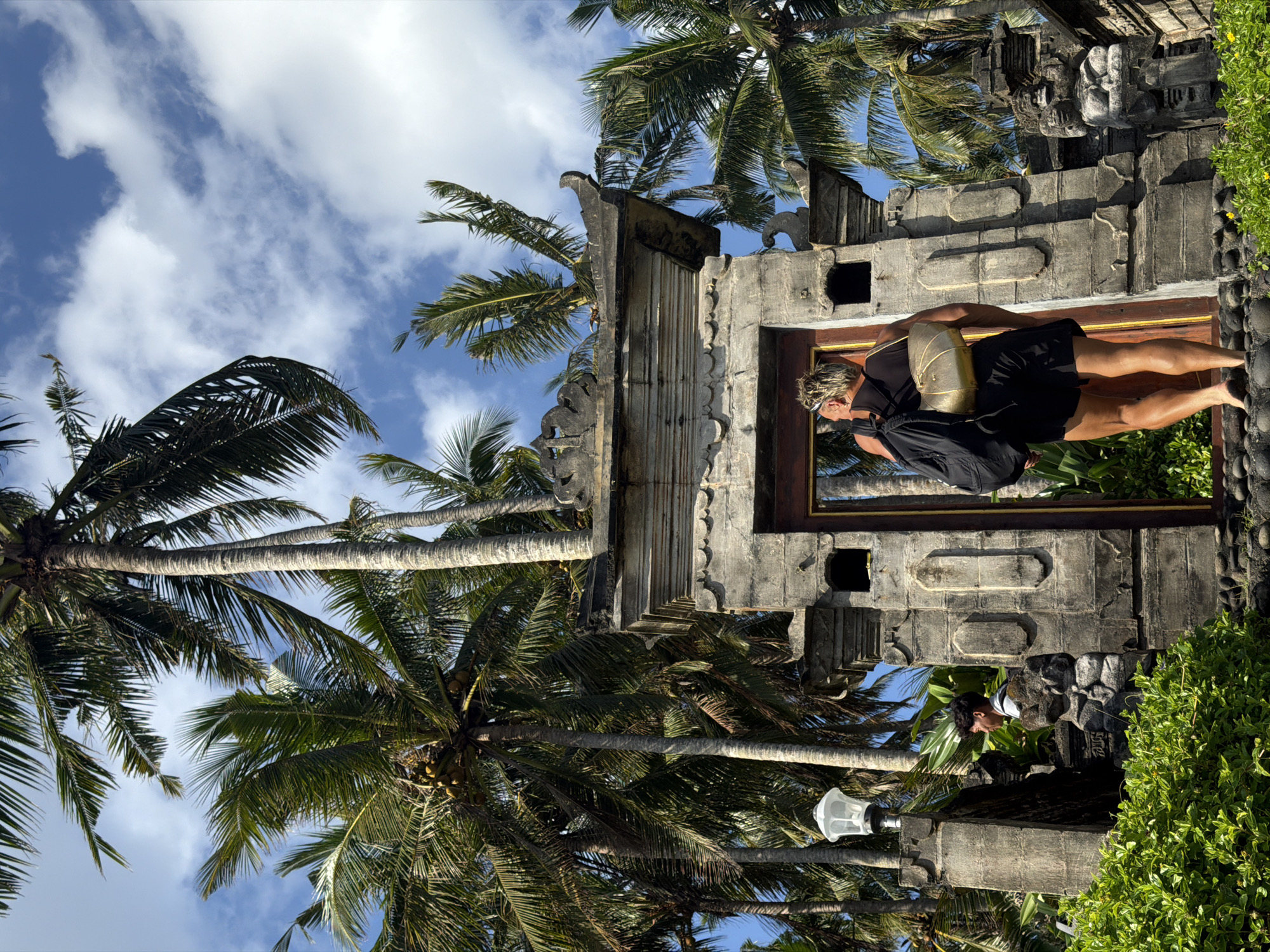A woman in a black tank top and black shorts stands in the doorway of a weathered stone structure. The doorway is framed in dark wood, and the structure itself has decorative stone carvings. Behind the structure and to the left are several tall palm trees with green fronds against a blue sky with scattered white clouds. To the right of the structure is dense green foliage. Another person is visible outside of the frame of the structure, at the bottom. Based on the architecture and environment, the location is likely Kerambitan, Indonesia.