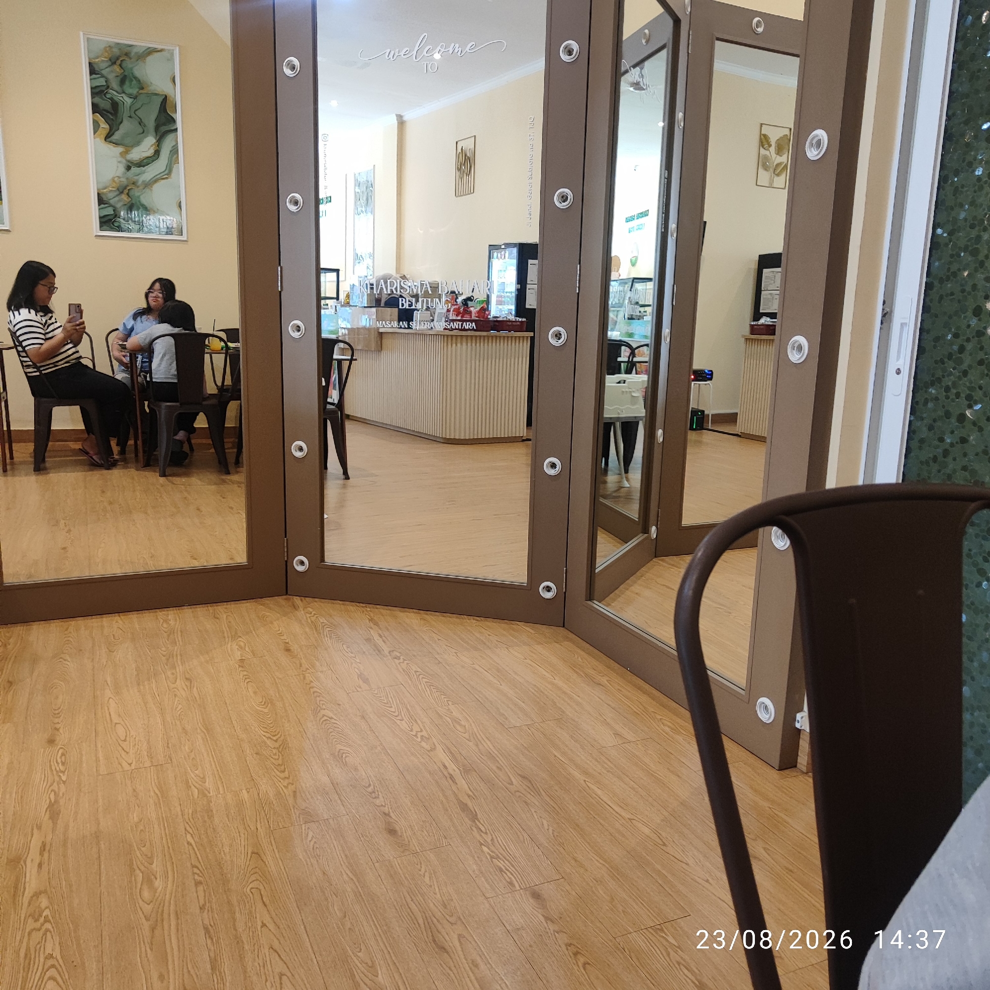 The image shows the interior of a cafe with a wooden floor. In the foreground, a dark brown chair is partially visible on the right. In the background, through a mirrored doorway, three people are seated at a table. A woman in a striped shirt is looking at her phone, and two other individuals, one wearing a blue shirt, are also seated. Behind them, a counter with the text "KHARISMA BELITUNG" is visible, along with a refrigerator and display cases. The cafe appears to be located in Tanjung Pendam, Indonesia, as indicated by the text on the counter. The overall scene suggests a casual dining or social setting.