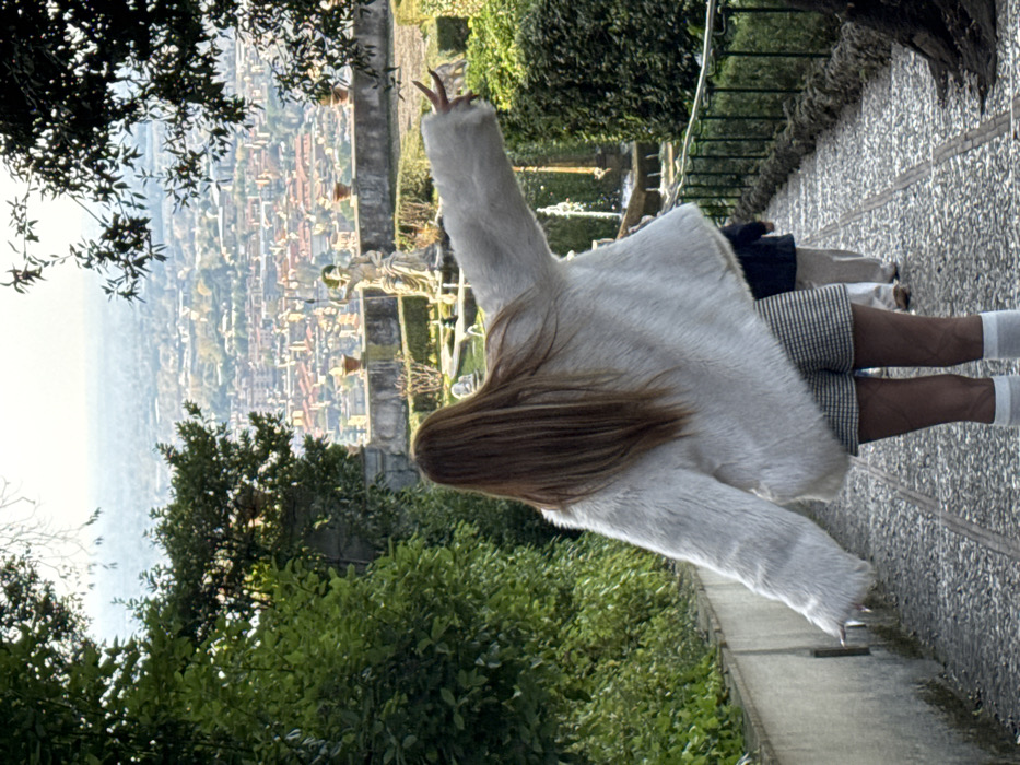 An individual, viewed from behind, stands on a cobblestone path within a terraced garden in Tivoli, Italy. The subject wears a white faux fur jacket, black and white plaid shorts, patterned dark hosiery, and white knee-high socks. Long, light brown hair extends down their back. The left arm is extended outwards, with fingers splayed. Two additional persons are visible further down the path, blurred and receding from the viewer. The immediate environment features dense green foliage flanking the path, a stone balustrade, potted plants, a classical statue depicting a male figure, and a functional fountain. Beyond the garden, a panoramic cityscape with numerous buildings, predominantly featuring terracotta roofs, extends to a hazy horizon with distant hills. A dark green metal railing lines the right side of the path.
