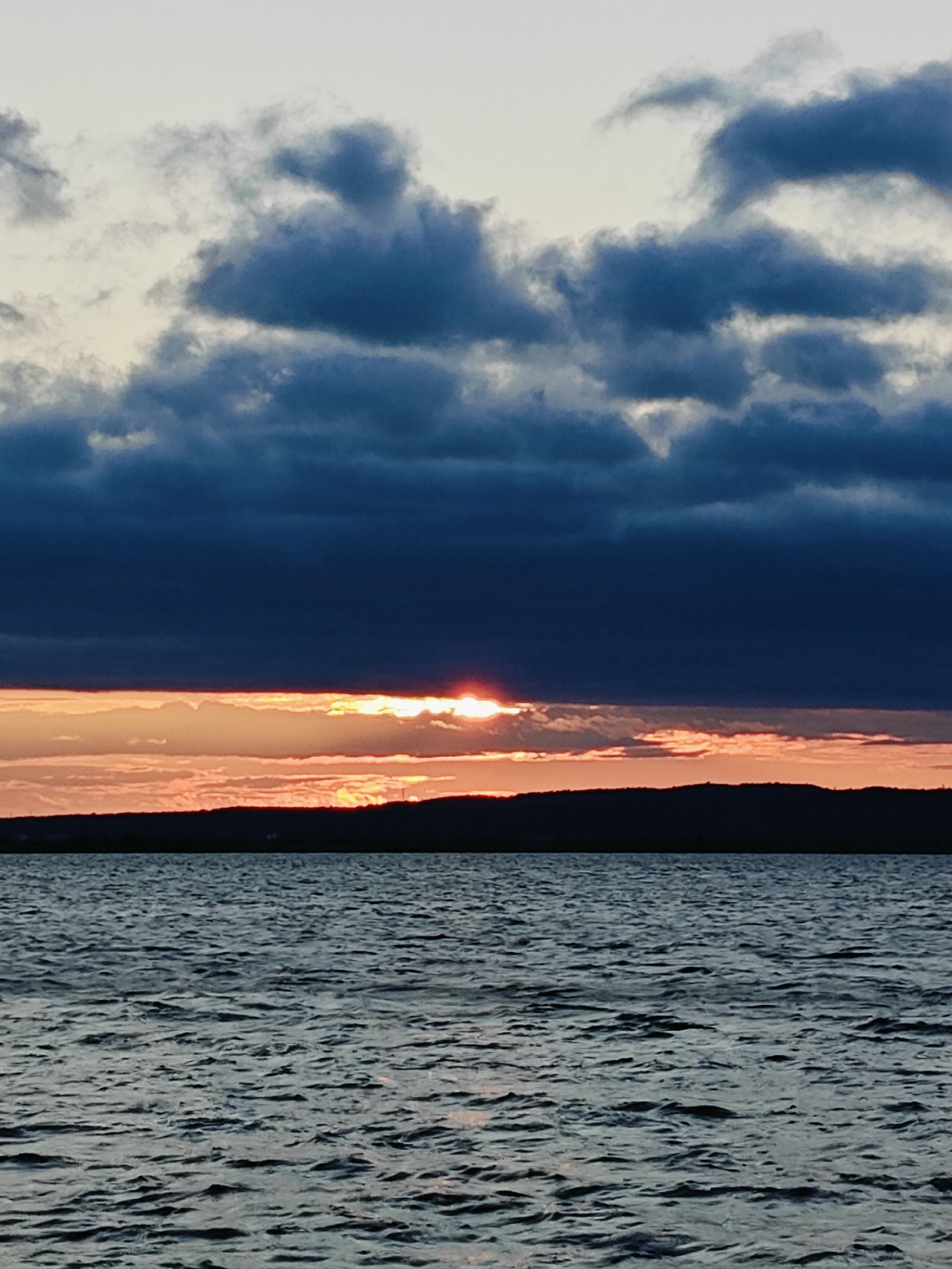 This is a vertical, eye-level, full-shot photograph of a sunset over a body of water, likely the Kama River in Chaikovsky, Russia. The sky is filled with dramatic, dark clouds that are illuminated from below by the setting sun. The sun itself is a small, bright sliver peeking through a gap in the clouds, casting an orange glow across the horizon. Below the sky is a dark silhouette of a distant, undulating shoreline, possibly with some sparse structures visible. The foreground is dominated by the rippling surface of dark water, reflecting the subdued light of the twilight sky. No people or specific objects are clearly discernible, emphasizing the natural landscape and the atmospheric conditions of the sunset.