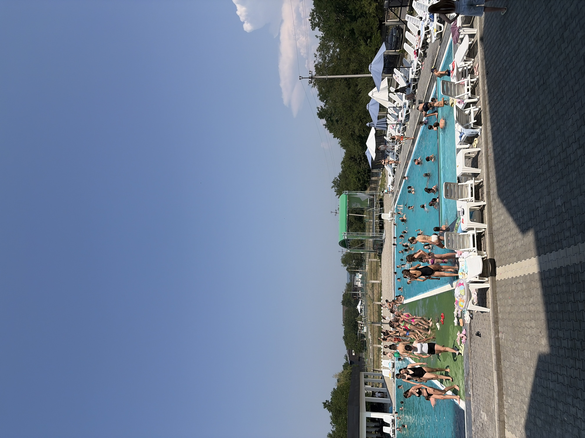 This image depicts a busy outdoor swimming pool complex on a sunny day, likely in Solotvyno, Ukraine. The pool itself is large and filled with numerous people of various ages, many of whom are swimming, splashing, and socializing. Several individuals are also standing on the pool deck, either entering or exiting the water, or relaxing.
Around the pool, numerous lounge chairs are set up, some occupied by people, while others are empty. Several umbrellas provide shade. The pool area is bordered by a paved walkway and beyond that, a grassy area with trees and some buildings. In the background, there are more trees and distant structures, suggesting a semi-rural or suburban setting. A green covered structure with a slide is visible on the far side of the pool. The sky is clear and blue, with a few small clouds. The overall atmosphere is one of recreation and leisure.