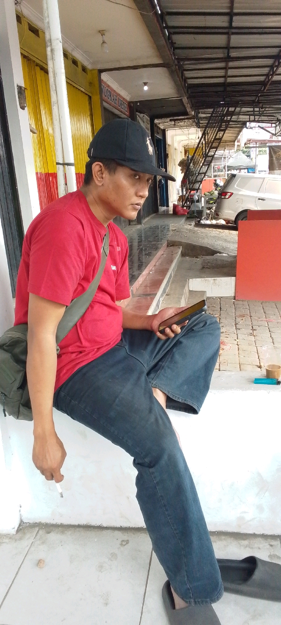 A man is sitting on a ledge in what appears to be an outdoor commercial area in Cilaku, Indonesia. He is wearing a red t-shirt, blue jeans, a black baseball cap, and a green messenger bag slung across his chest. He is holding a smartphone in his right hand and a lit cigarette in his left hand. The background shows a covered walkway with a metal roof supported by columns. A yellow and red corrugated metal shutter is visible on the left, and a white building with a tiled floor is on the right. A white car is parked in the background, along with stairs and other structures. There is a plastic cup with a beverage, a blue lighter, and a roll of tape on the ledge near the man.