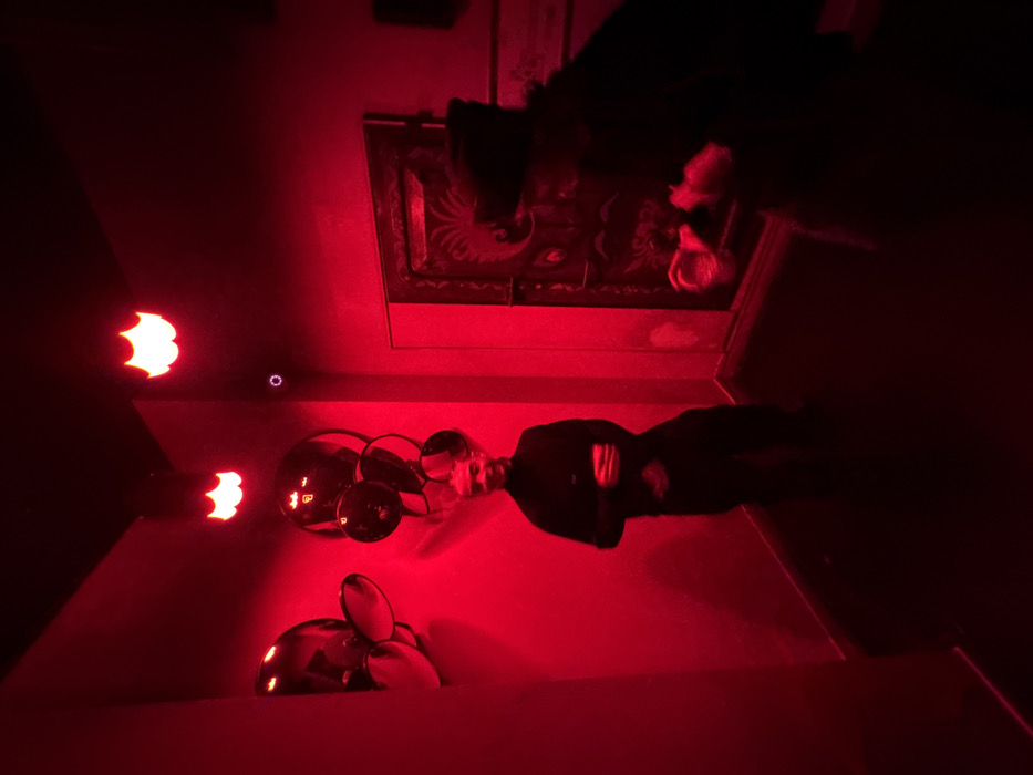 A red-lit room featuring a man with crossed arms standing on a floor. Decorative items are positioned on the floor, appearing to be glass objects. Above, a framed artwork or design hangs on the wall. The scene is illuminated by an intense red light, creating a strong contrast and obscuring details. Lights on the wall provide additional red illumination. A woman is standing in the frame.